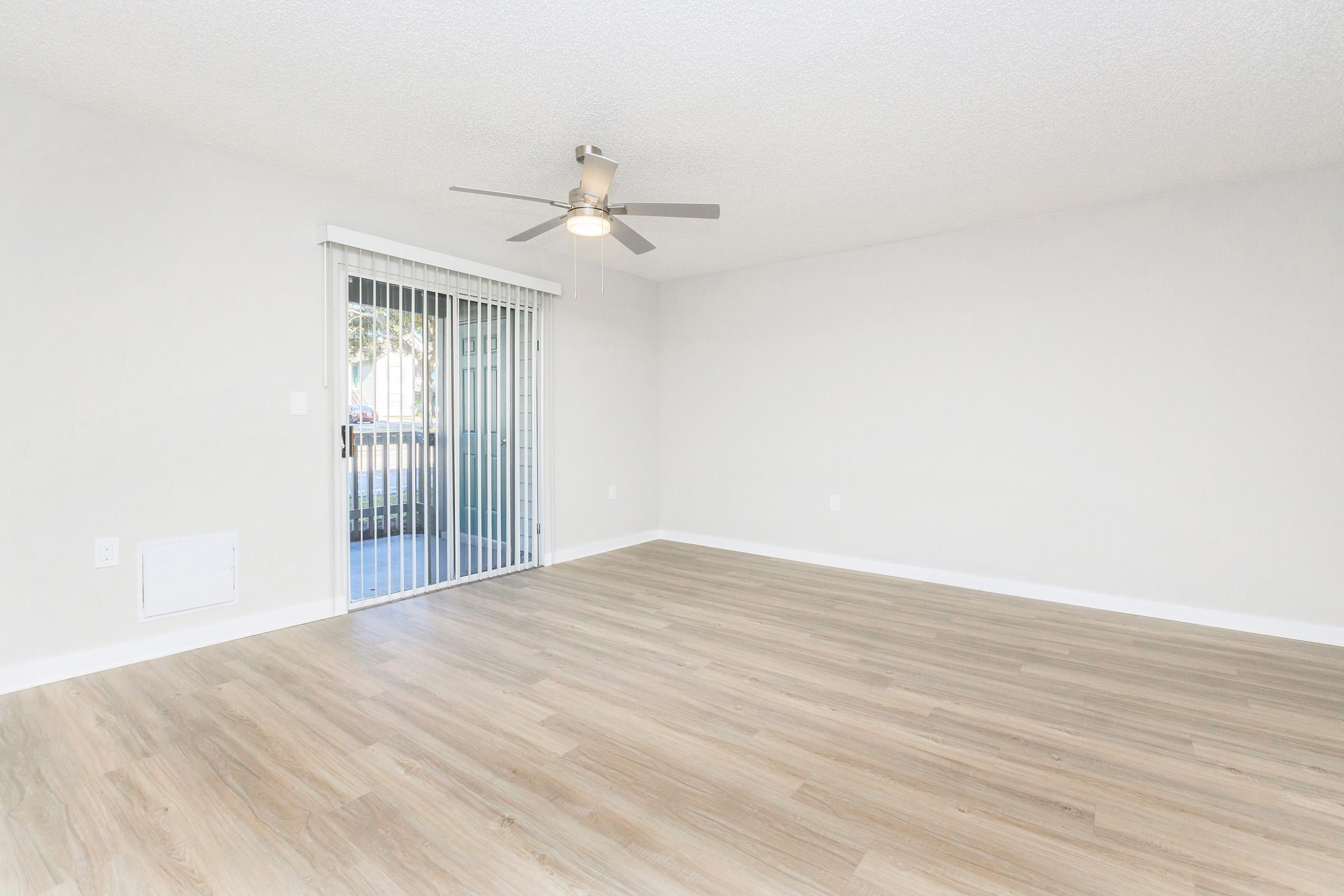 A spacious, empty living room with light wooden flooring and a ceiling fan. A sliding glass door leads to an outdoor area, allowing natural light to enter. The walls are painted in a neutral color, creating a bright and airy atmosphere.