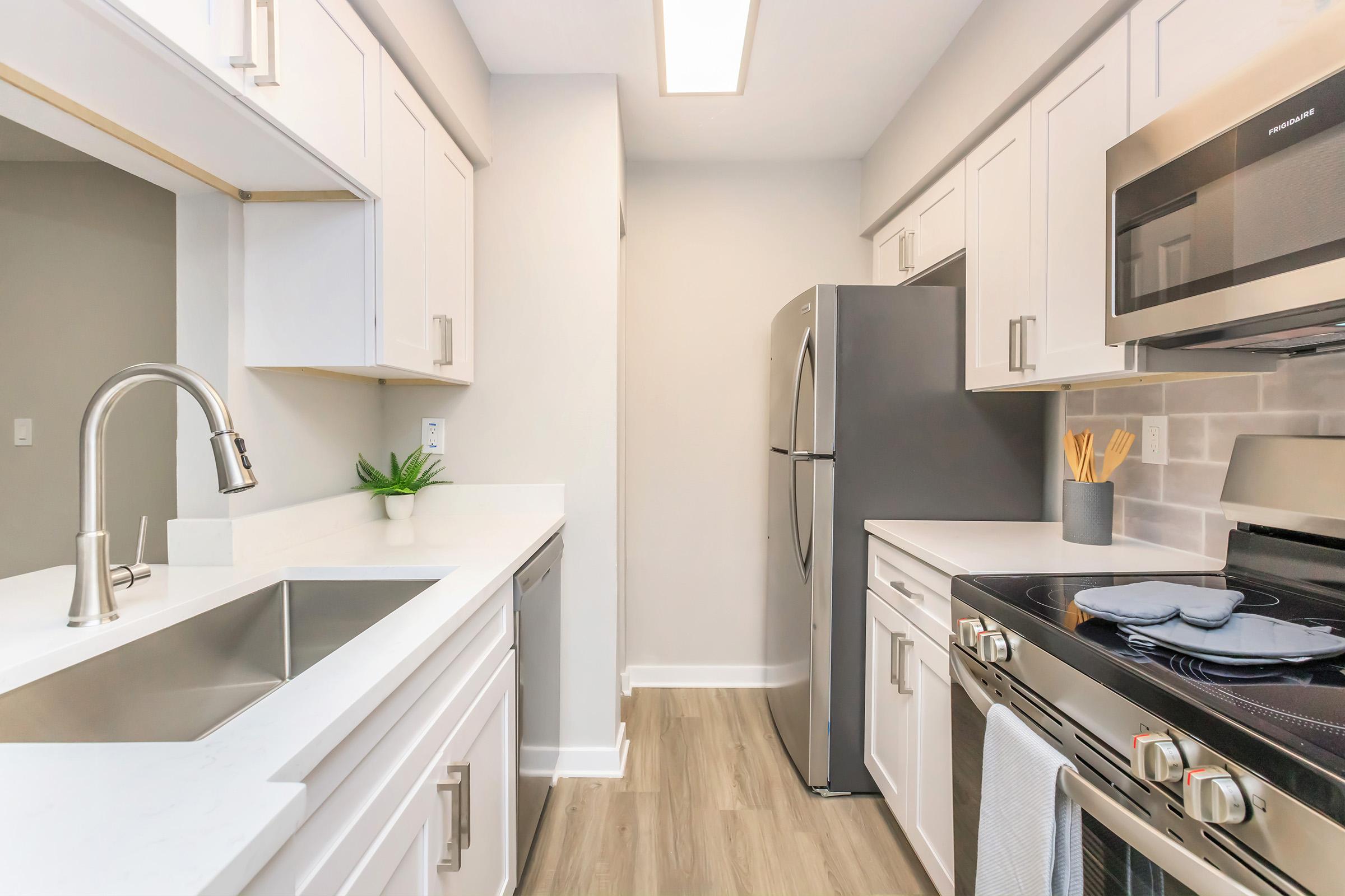 Modern kitchen featuring white cabinets, stainless steel appliances, a double sink, and a gray refrigerator. The countertops are light-colored, and a small plant adds a touch of greenery. The room is well-lit, with a neutral color scheme and a clean, organized appearance.
