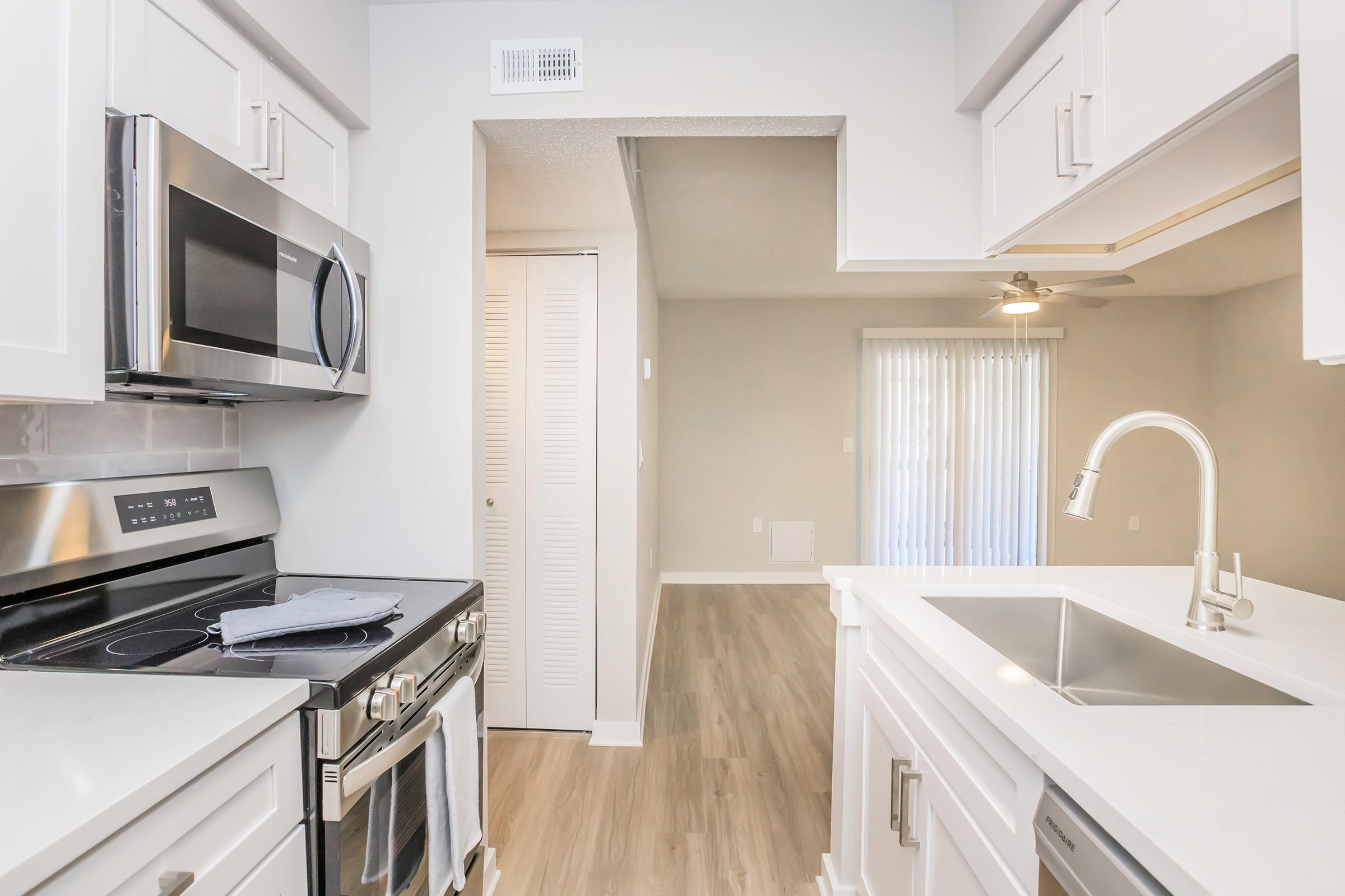 A modern kitchen featuring stainless steel appliances, including a microwave and stove, white cabinetry, and a sink with a sleek faucet. Light-colored walls and flooring create a spacious feel. In the background, there is a living area with sliding glass doors and a ceiling fan, providing ample natural light.