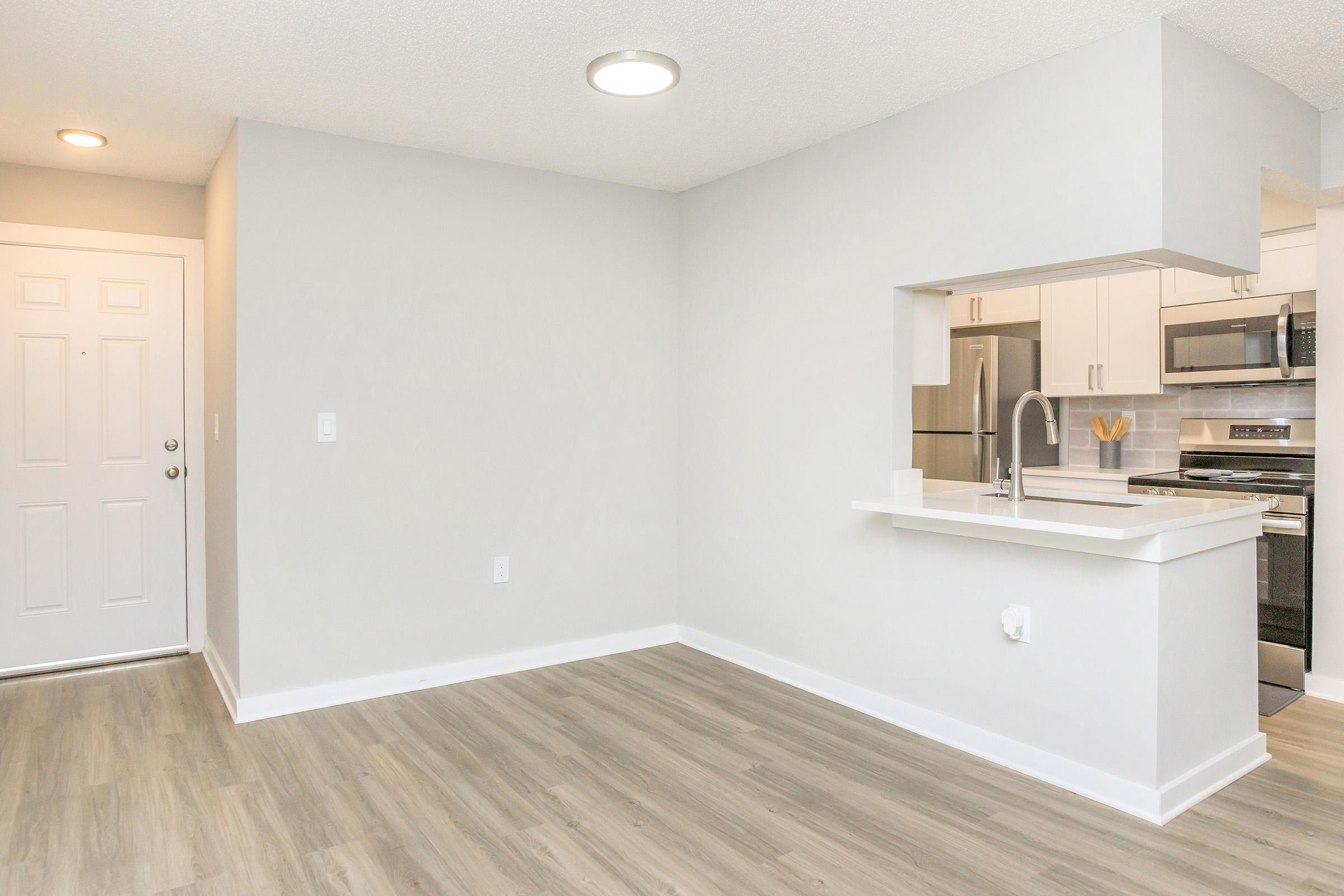 A modern, minimalist living space featuring light gray walls and a light wood floor. There’s a small kitchen area with a white countertop and stainless steel appliances. A front door is visible on the left, and the room is well-lit with natural light, creating an inviting atmosphere.