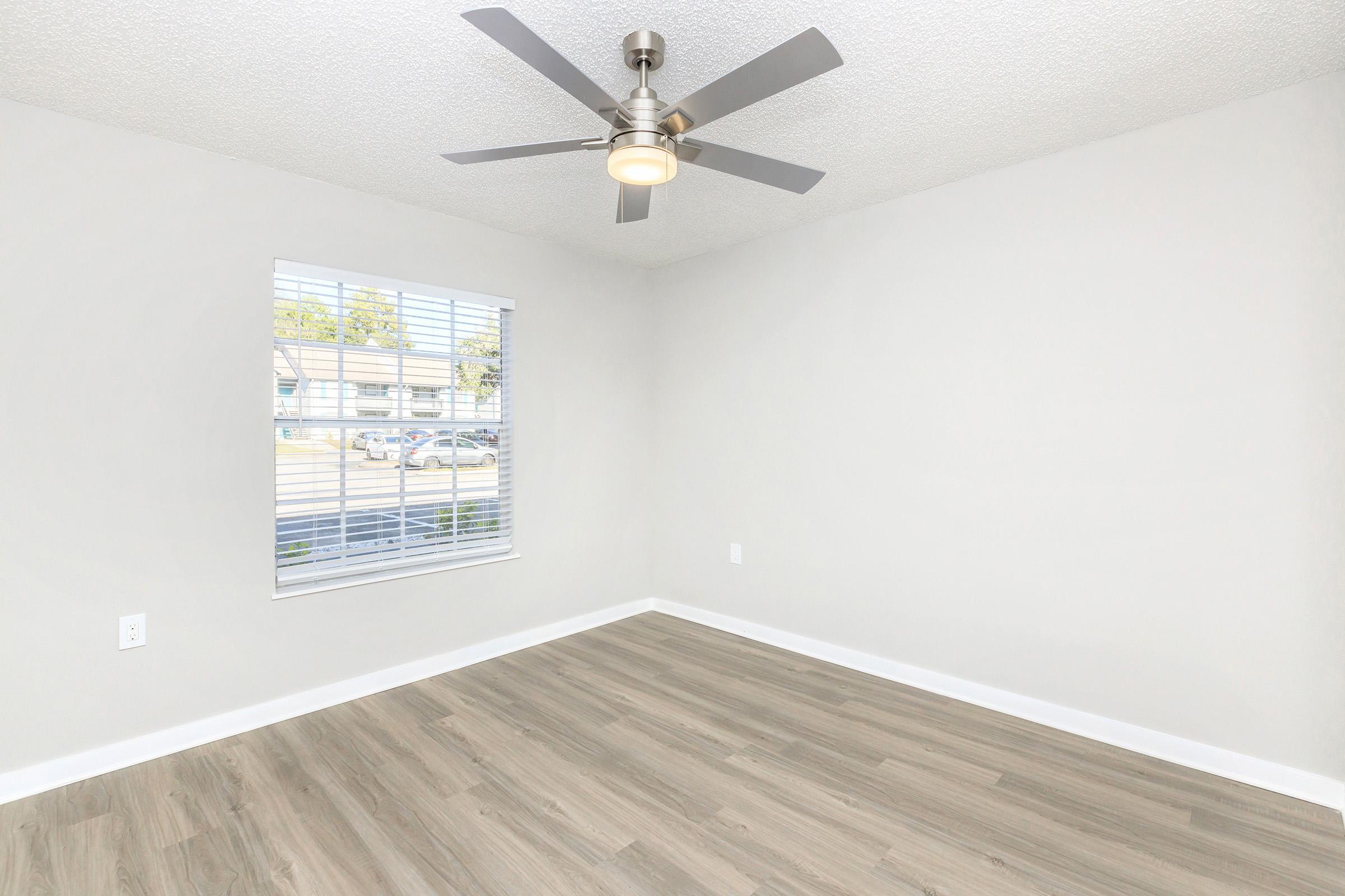 A bright, empty room with light gray walls and a light-colored wood floor. A ceiling fan with a light fixture hangs in the center, and a window with white blinds allows natural light to enter from outside, offering a view of parked cars and greenery.