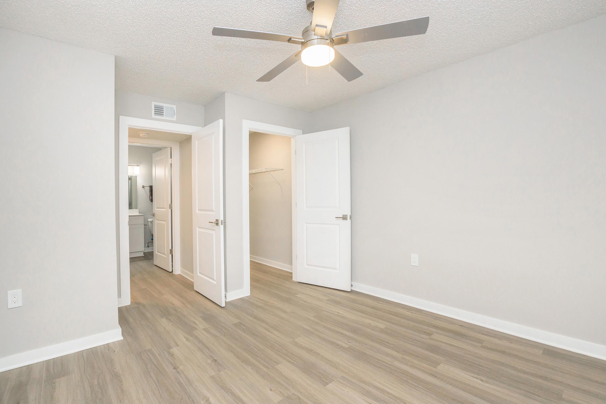 A spacious, light-colored room featuring a ceiling fan and laminate flooring. There are two white doors: one leading into a bathroom and the other into a closet. The walls are painted in a soft gray, creating a modern and inviting atmosphere. The room is empty, allowing for various furniture arrangements.