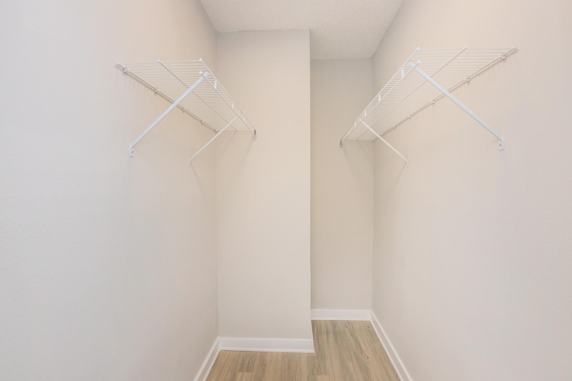Empty closet space with two wall-mounted wire shelves on opposite walls, light-colored walls, and a polished wooden floor. The space appears bright and clean, ideal for organizing clothing or storage items.
