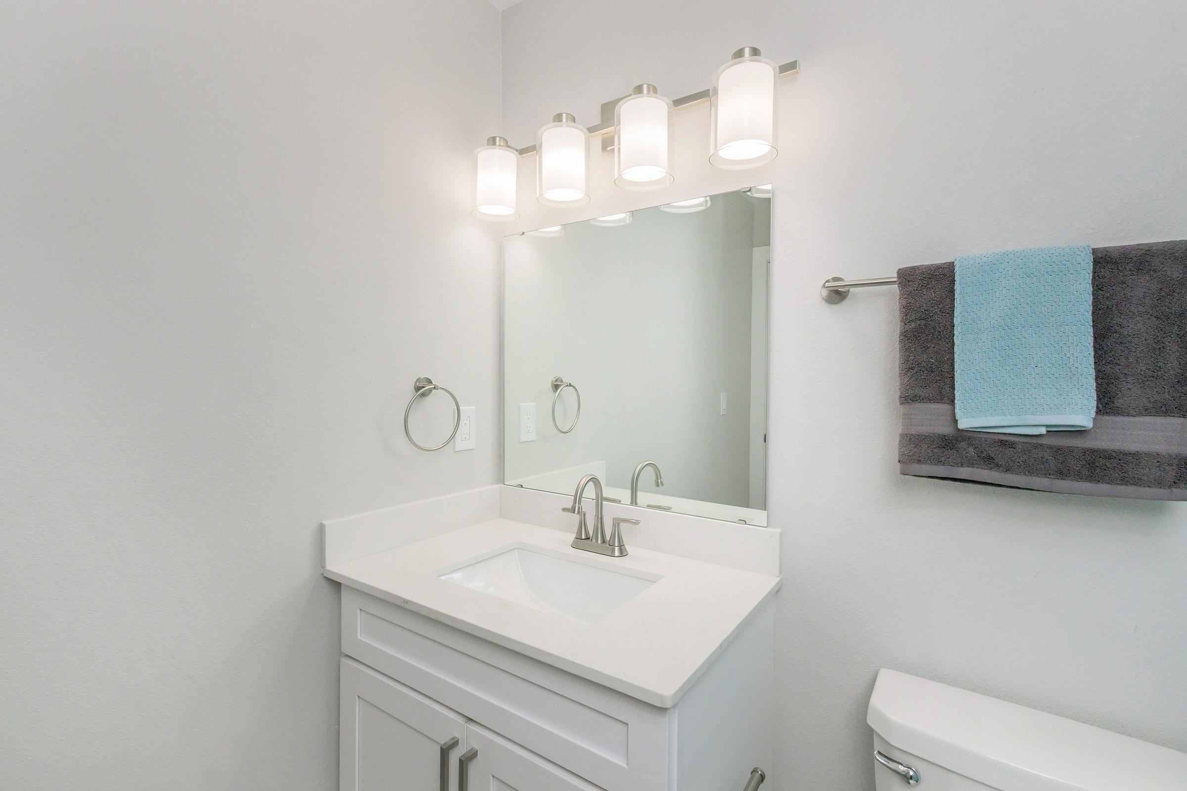 Modern bathroom with a white vanity and sink. A large mirror above the sink is illuminated by four light fixtures. A gray towel hangs on a rack, and a blue towel is neatly placed on the countertop. The walls are light-colored, creating a clean and bright atmosphere.