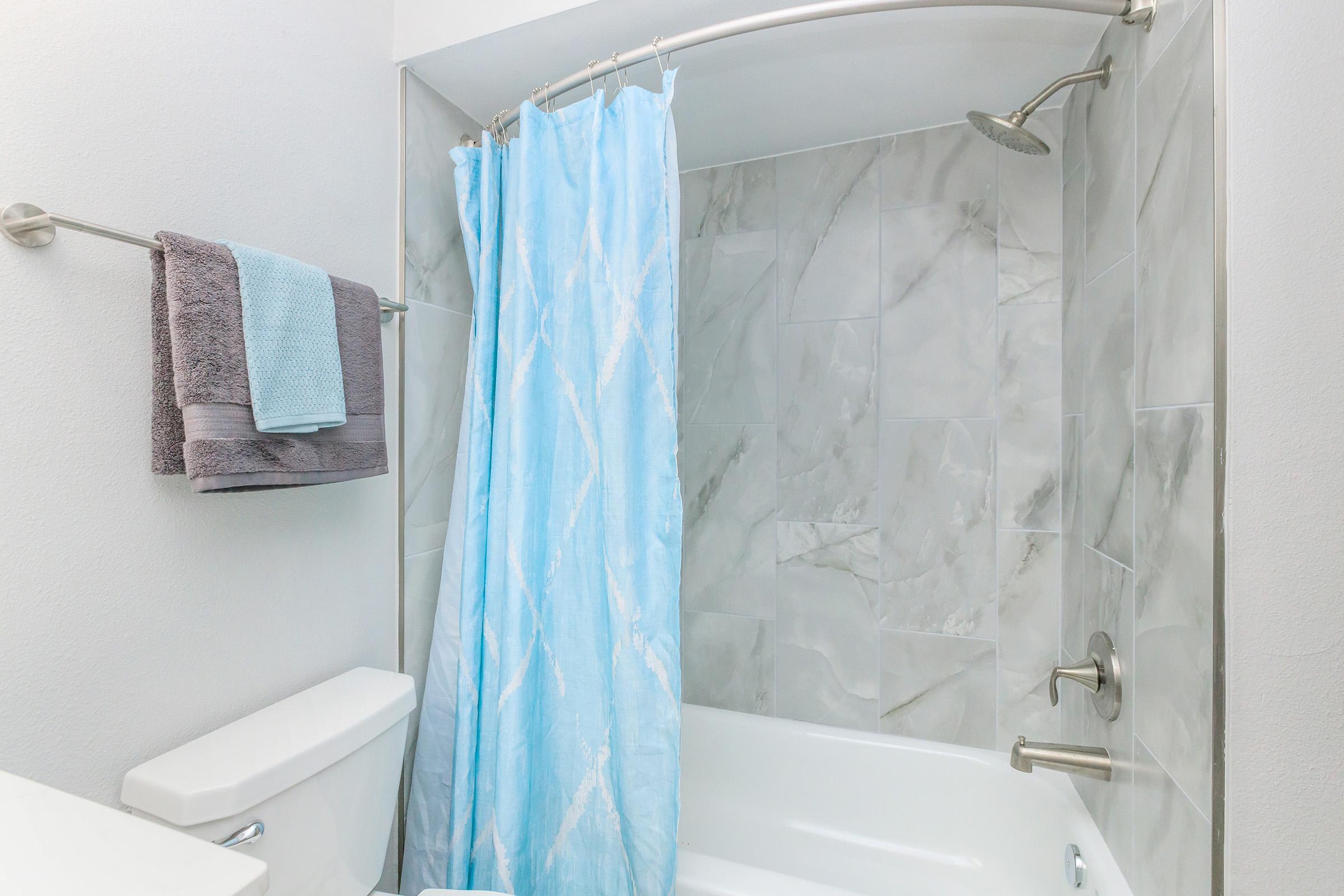 A clean bathroom with a shower featuring a blue curtain, light gray tiles, a silver showerhead, and neatly arranged towels in shades of gray and light blue hanging on a towel bar. The bathtub is white and complements the modern design.