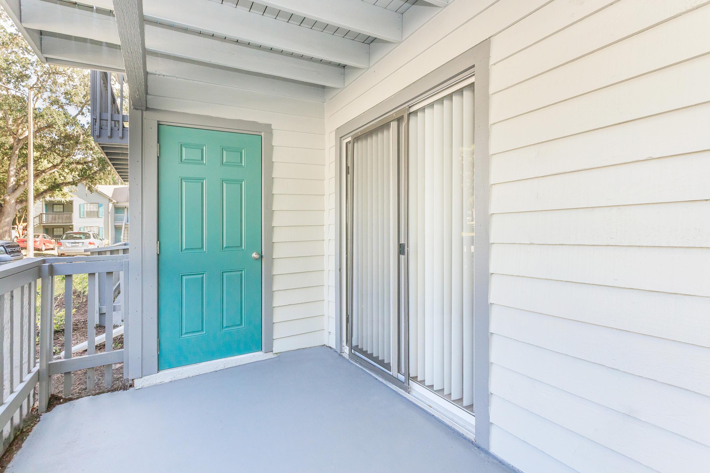 A porch area featuring a turquoise door and sliding glass doors with white vertical blinds. The walls are white wooden siding, and the floor is gray. The setting appears to be part of a residential building, with greenery visible in the background.