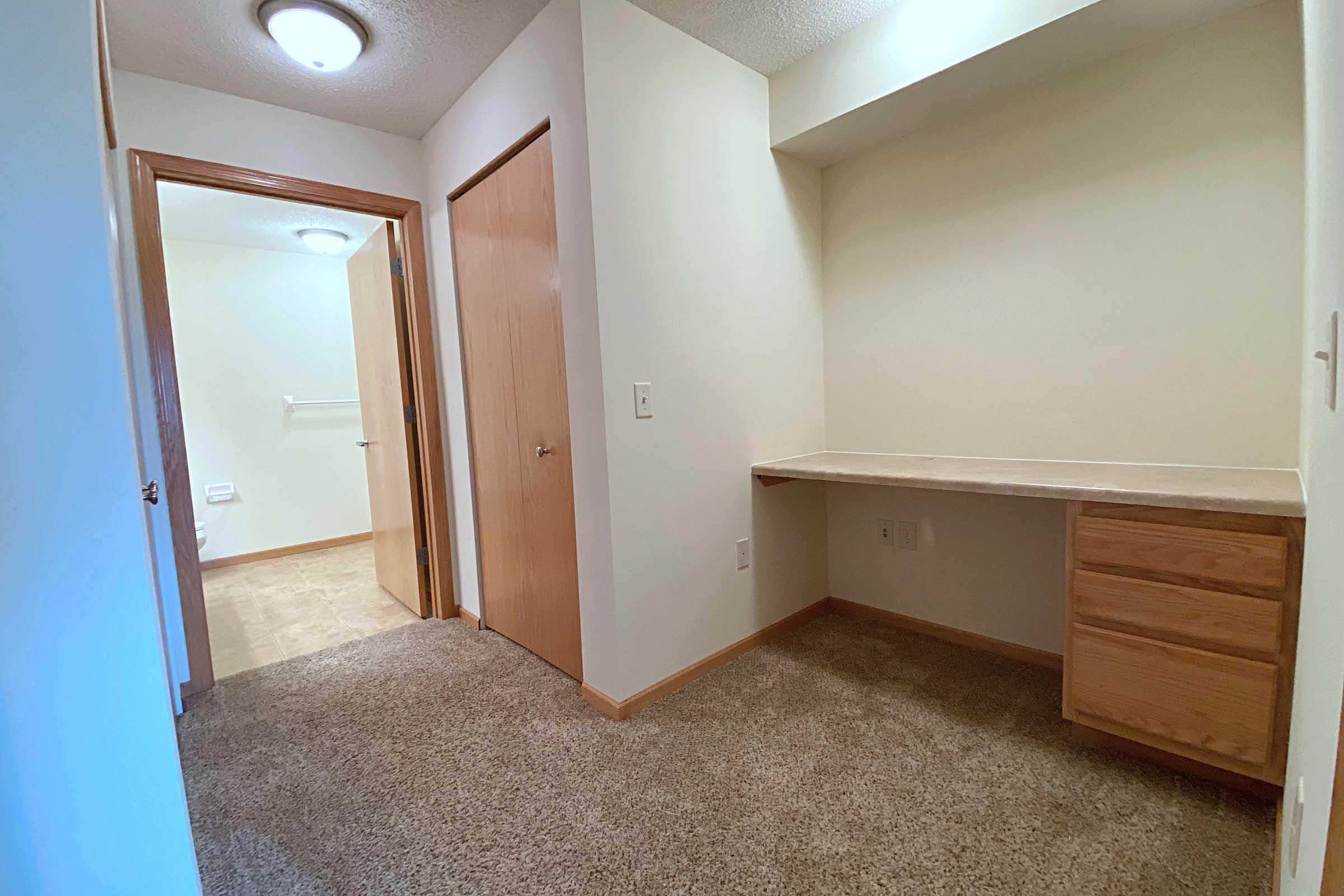 A hallway with tan carpet and light-colored walls. On one side, there is a small desk area with drawers. A door leads to another room, and there is a light fixture on the ceiling. The space is well-lit and has a clean, simple design.