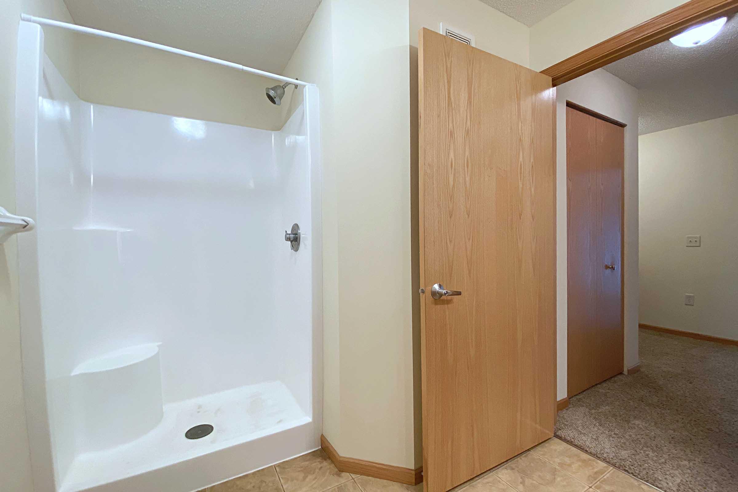 A bathroom interior featuring a white shower stall on the left, a wooden door on the right, and beige walls. The floor has light-colored tiles, and a carpeted area is visible in the adjacent room. Natural light illuminates the space.