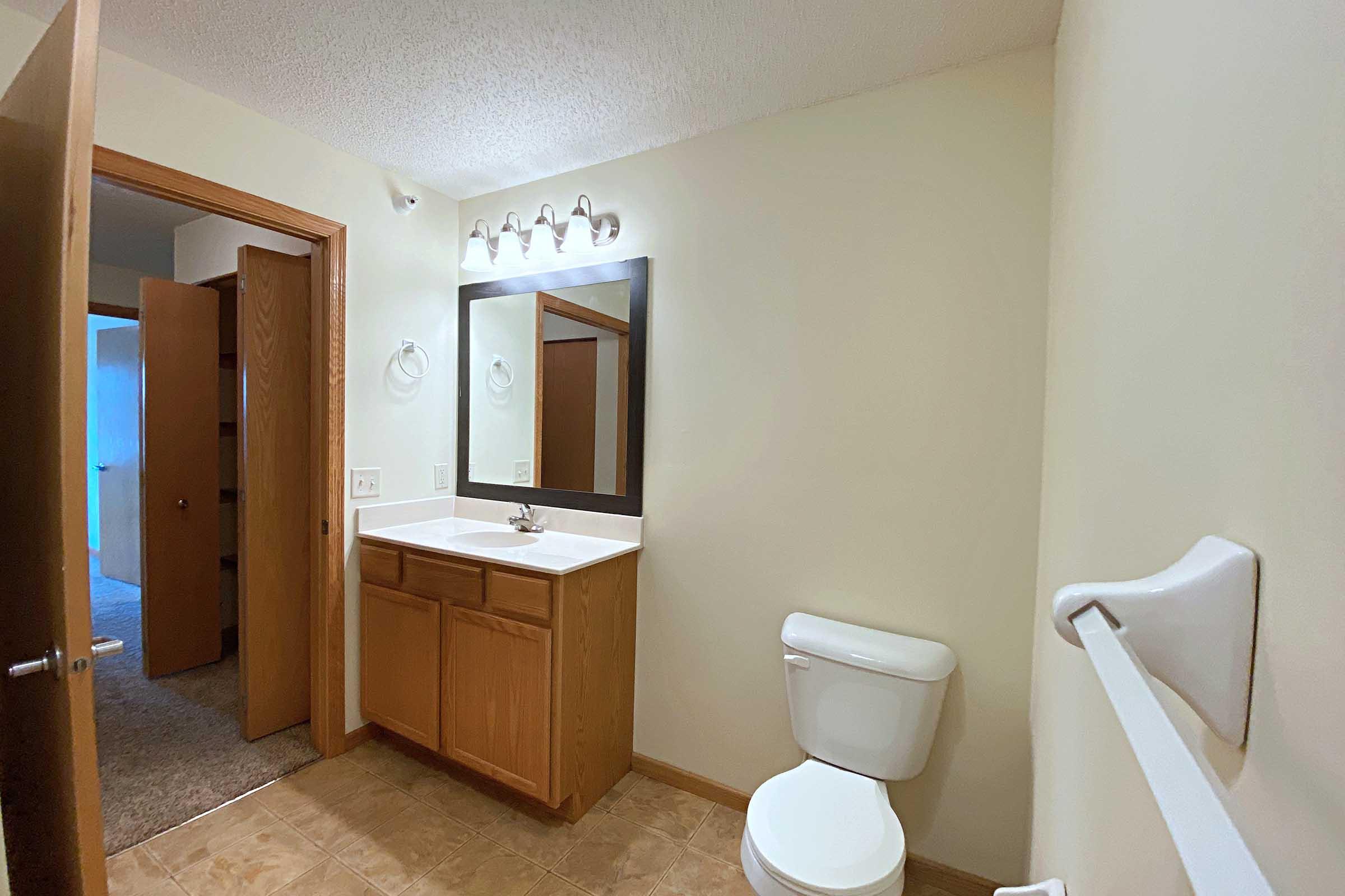 A bright bathroom featuring a sink with a wooden cabinet, a mirror above it, and a toilet. The room has beige walls and a tiled floor. A wooden door leads to another room, and there are towel rings mounted on the walls. The overall space is clean and well-lit.