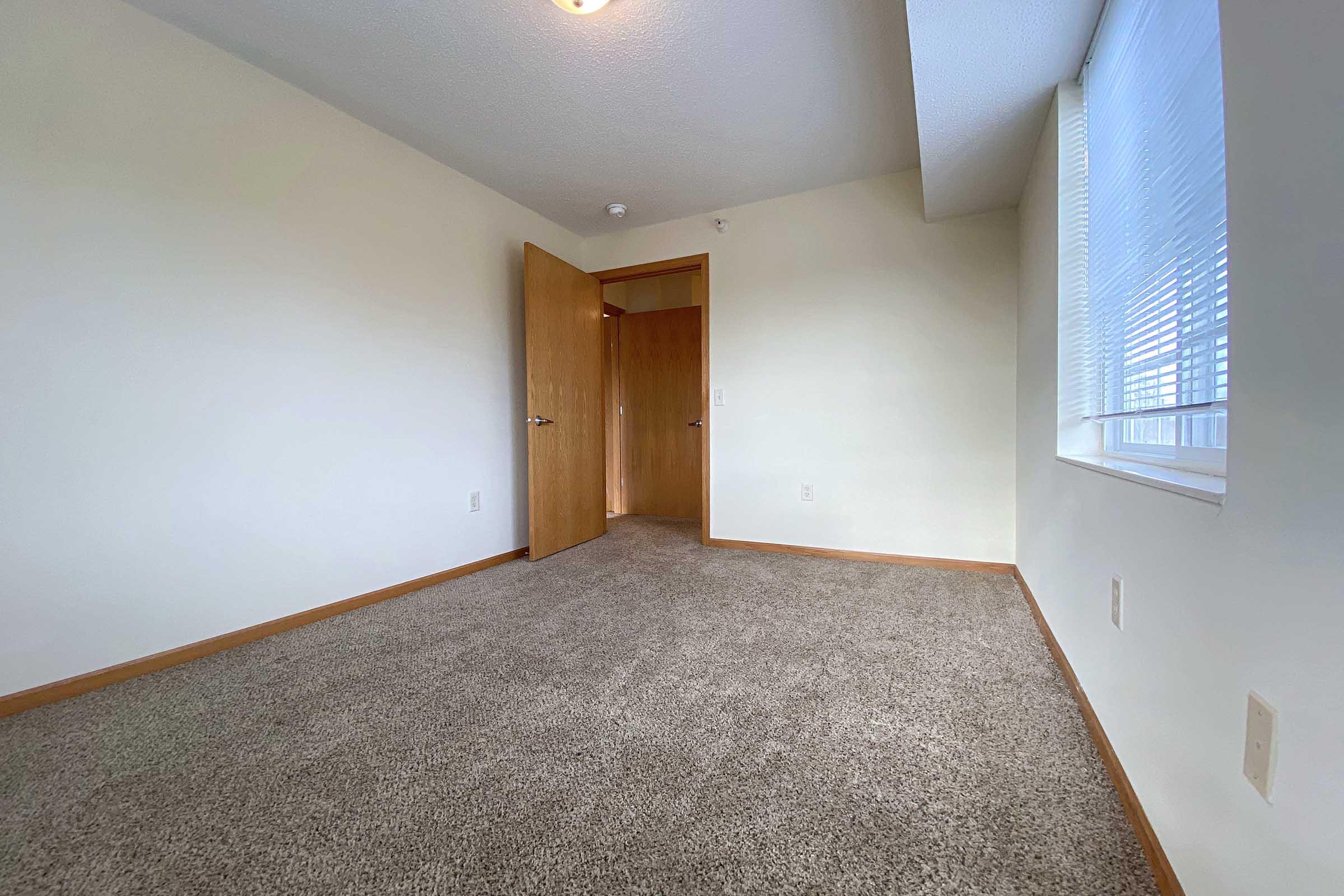 Empty room featuring light-colored walls, a carpeted floor, and a partially open door leading to another room. A window is visible with light coming in, creating a bright ambiance. The space is uncluttered and minimalistic, suitable for various uses.