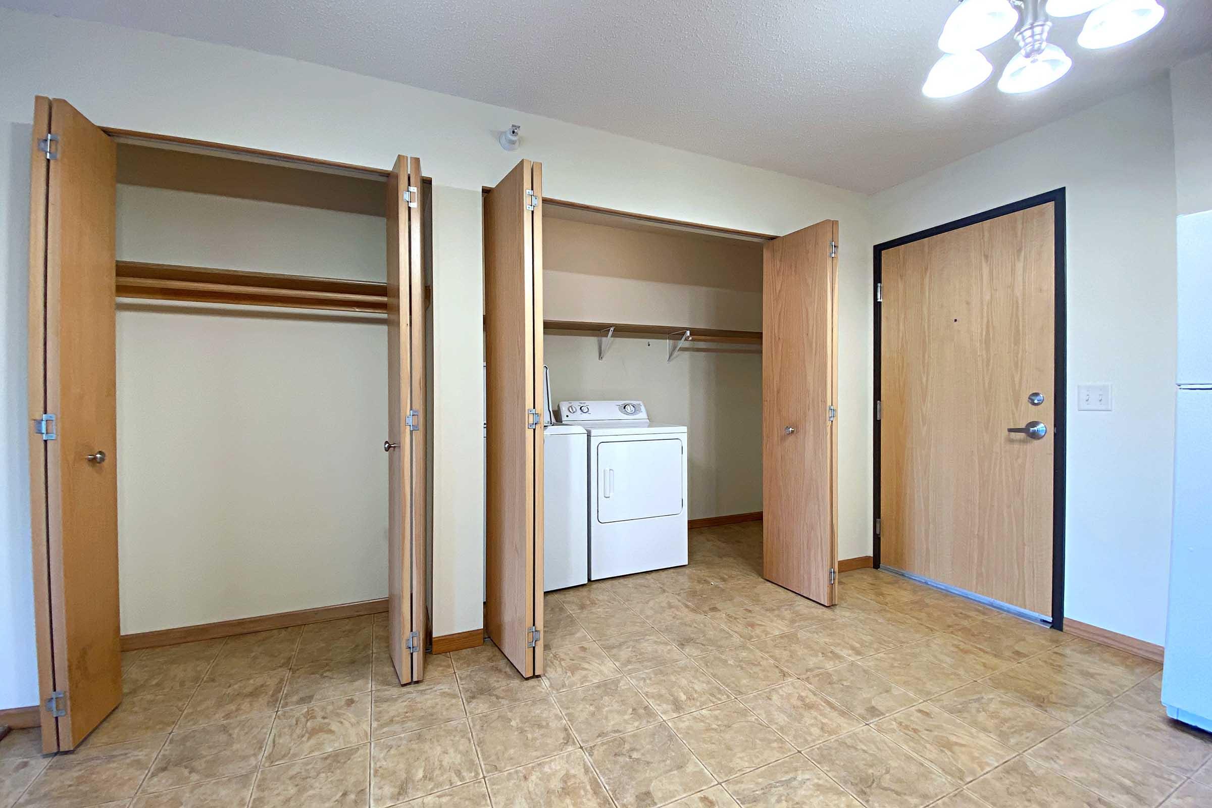 A laundry area featuring a washing machine and dryer, with two open closet doors on the left. The space has a light-colored tiled floor and a front door visible on the right. Overall, it appears clean and well-lit, suitable for household chores.