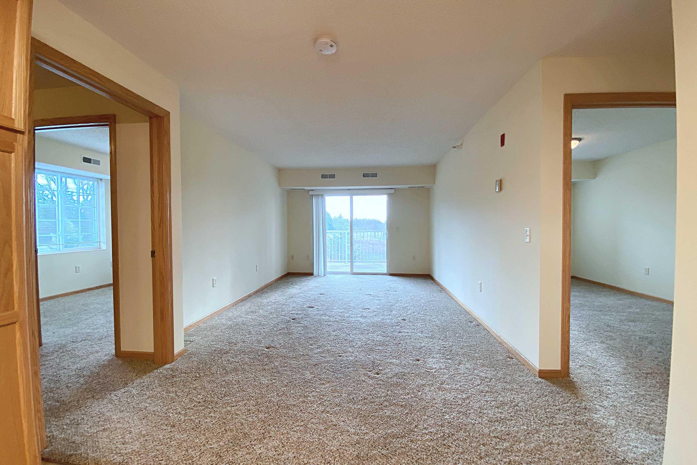 A spacious, well-lit living area featuring beige walls and carpet flooring. There are doorways leading to adjacent rooms on either side, and a large window with a view to the outside. The room is empty, providing an open feel, and the overall ambiance is neutral and inviting.