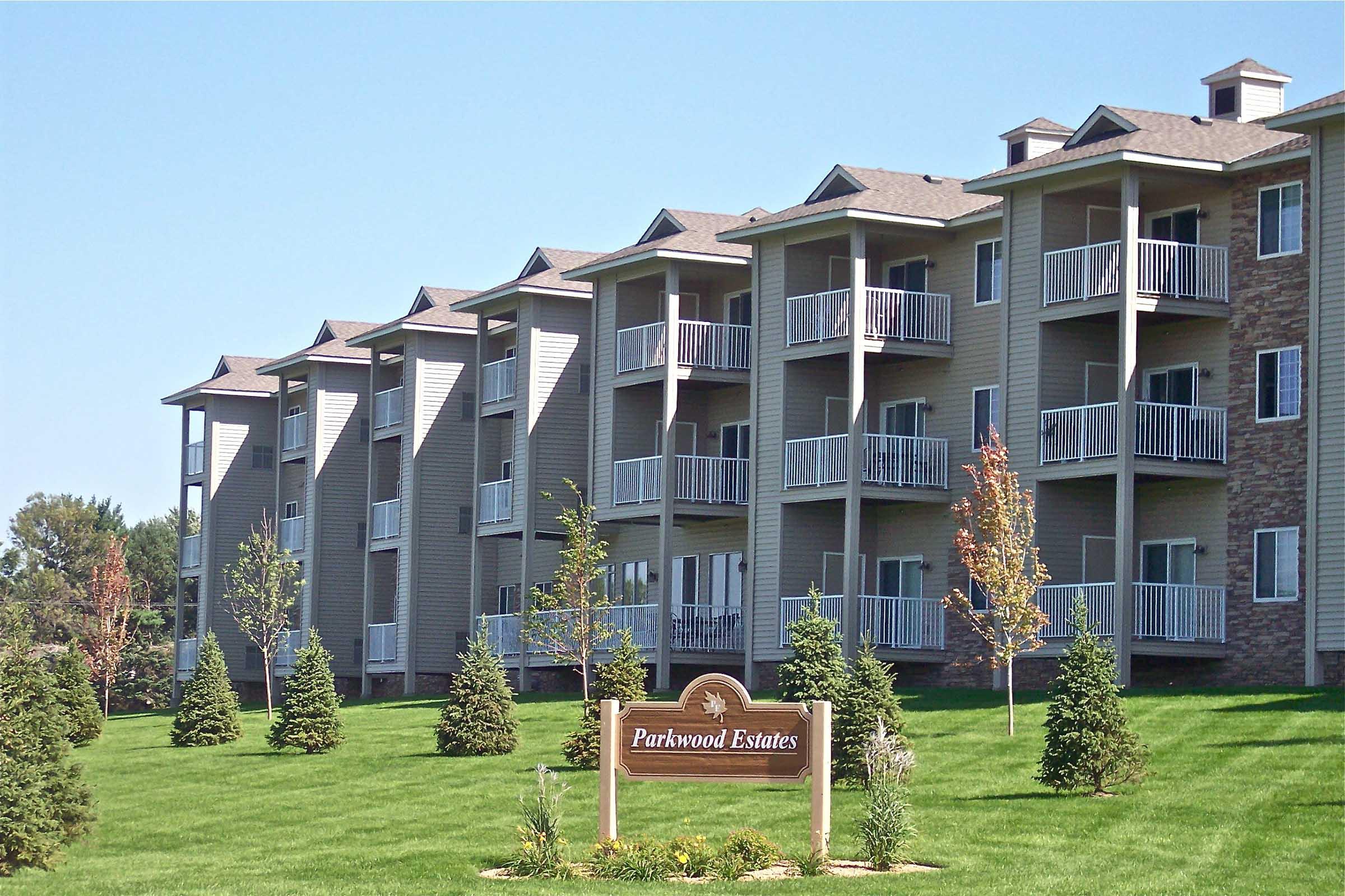 A row of modern, multi-story apartment buildings with balconies, set on green grass. A sign in front reads "Parkwood Estates," indicating the name of the complex. Small trees and landscaping are visible, and the sky is clear and sunny.