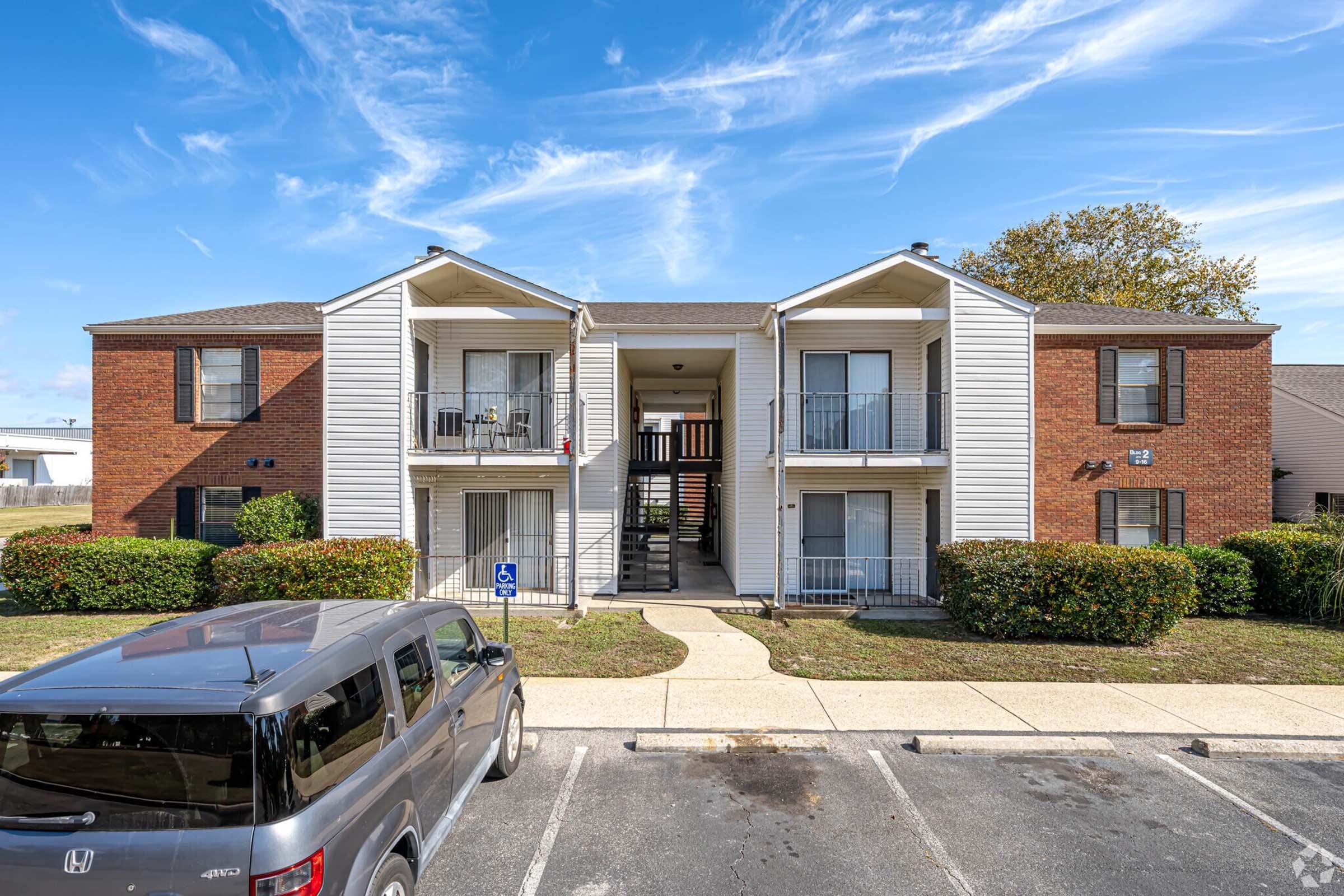 A two-story apartment building with a mix of brick and siding exteriors. Each unit has a small balcony and is surrounded by neatly trimmed bushes. A gray SUV is parked in the foreground, and a blue sky with wispy clouds is visible above.