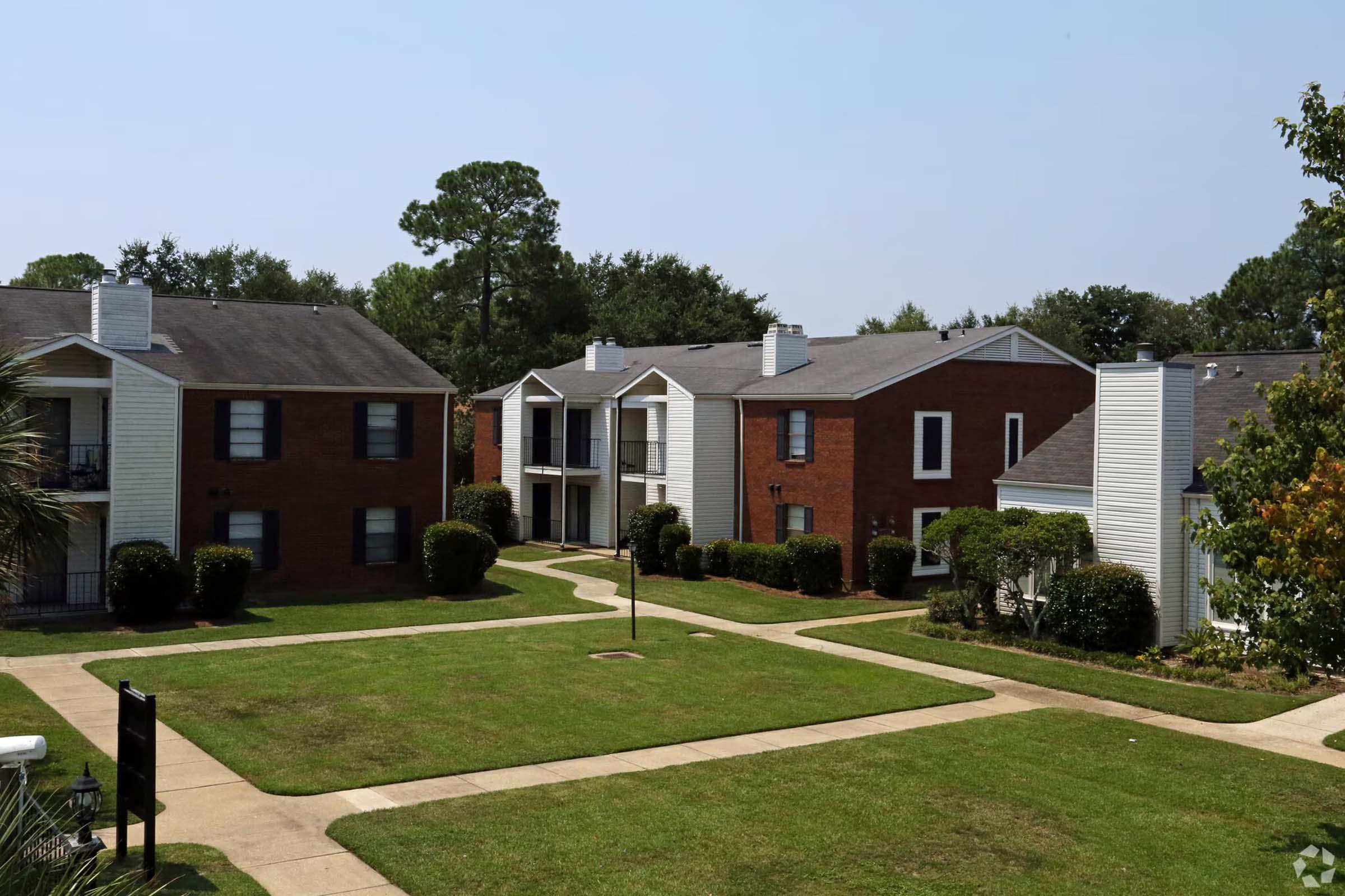 A view of an apartment complex featuring red brick and white exterior buildings, surrounded by neatly manicured lawns and shrubs. The layout includes walkways connecting the buildings, with trees in the background and a clear blue sky above.