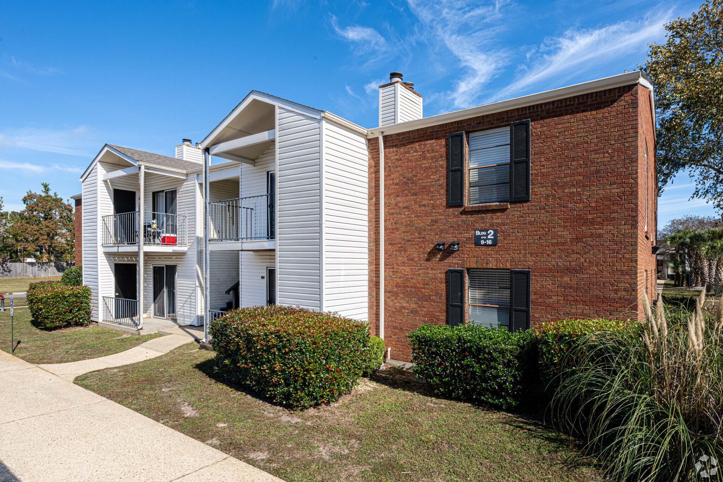 Two-story apartment complex featuring a mix of white siding and brick exterior. The left side has balconies, while the right side is brick with windows. A pathway leads up to the buildings, surrounded by shrubs and grass, under a clear blue sky. Street signage indicates "Building 2" is present.