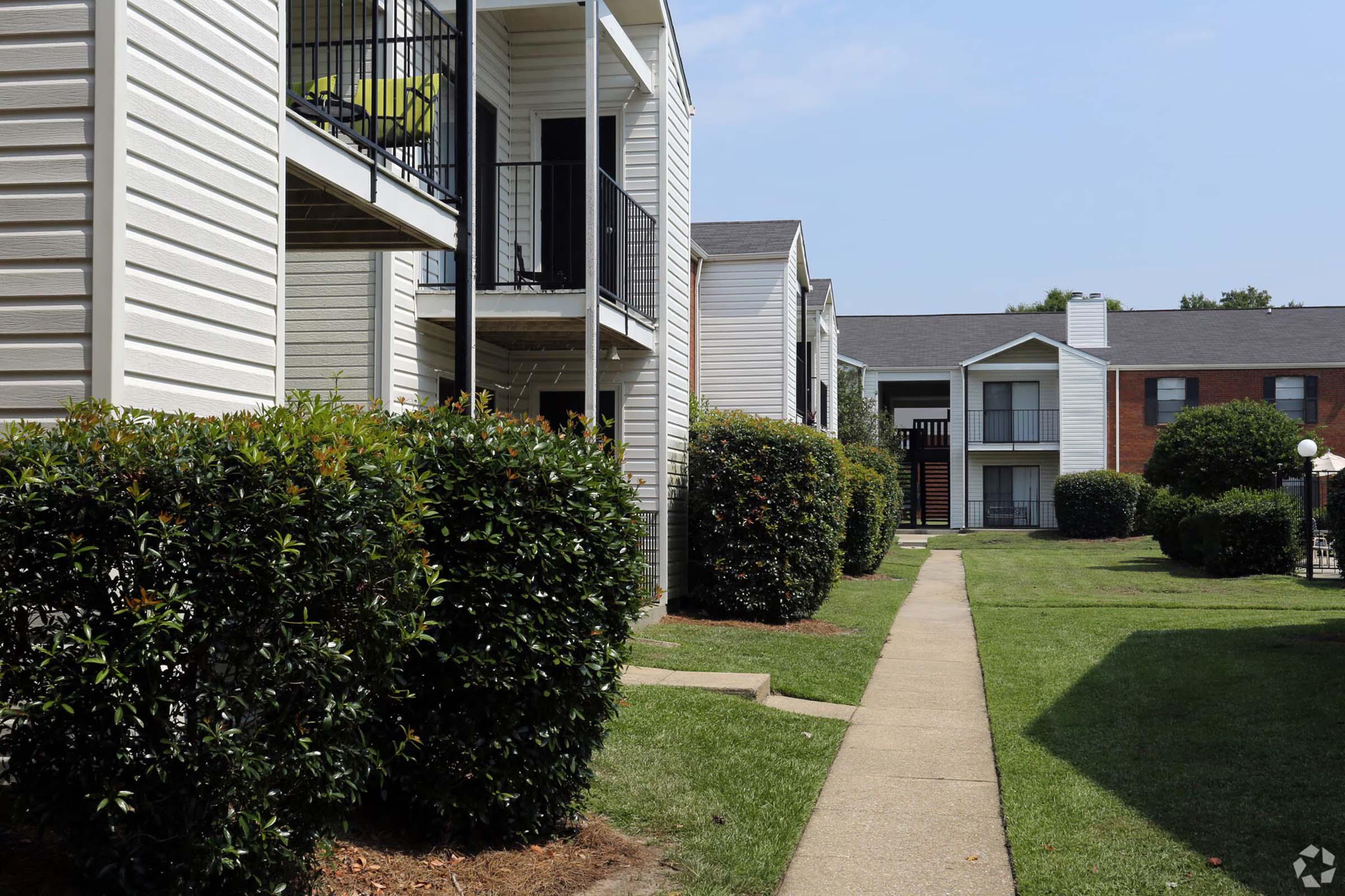 Row of apartment buildings with white siding and balconies, separated by well-maintained hedges and a grassy walkway. The scene is well-lit, showcasing a clear blue sky and a clean, inviting outdoor environment.