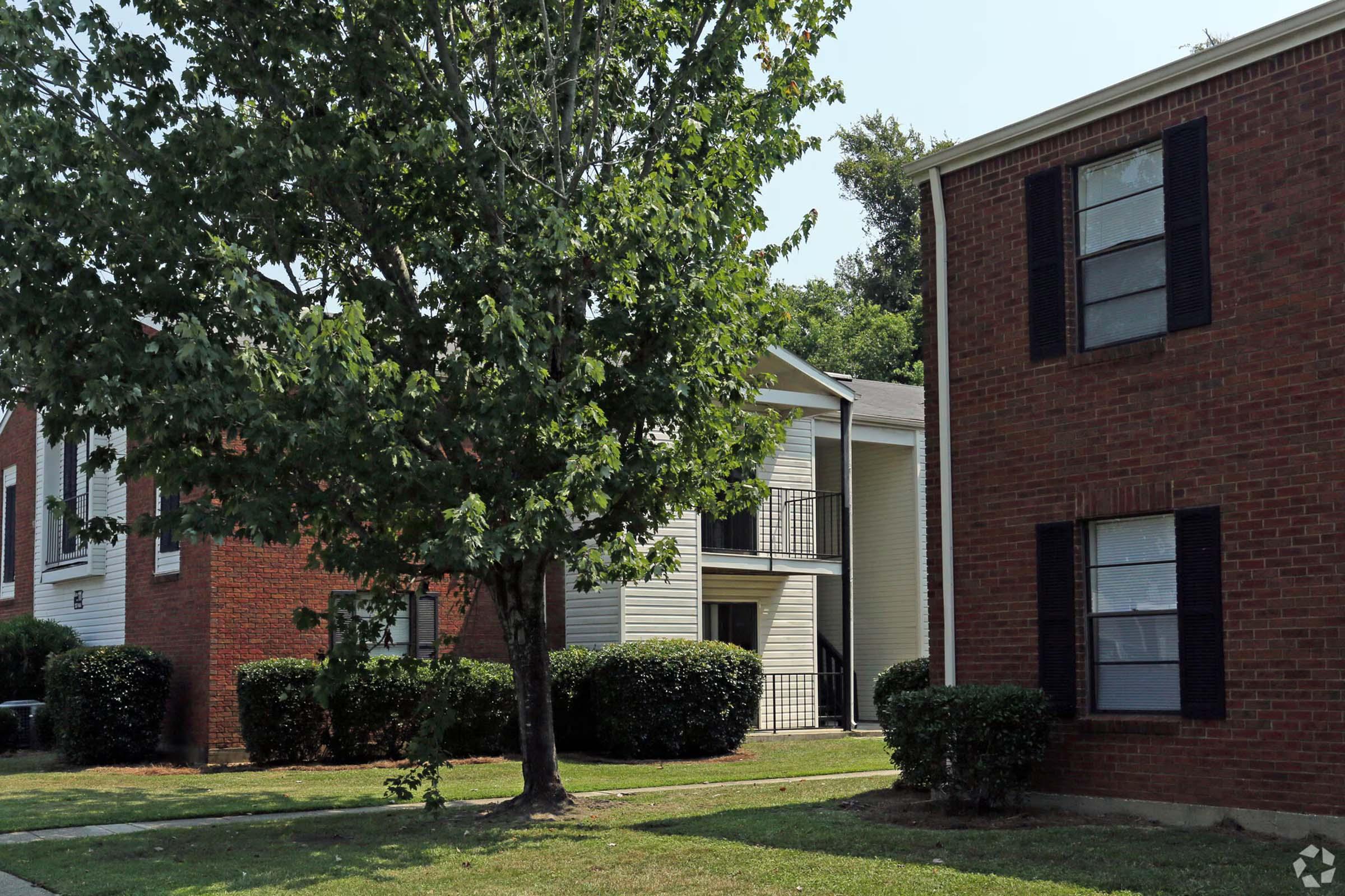 A residential area featuring two multi-story apartment buildings. The building on the right is made of red brick with black shutters, while the building on the left has a light-colored exterior with a balcony. A large tree and trimmed bushes are visible in the foreground, along with a grass lawn.