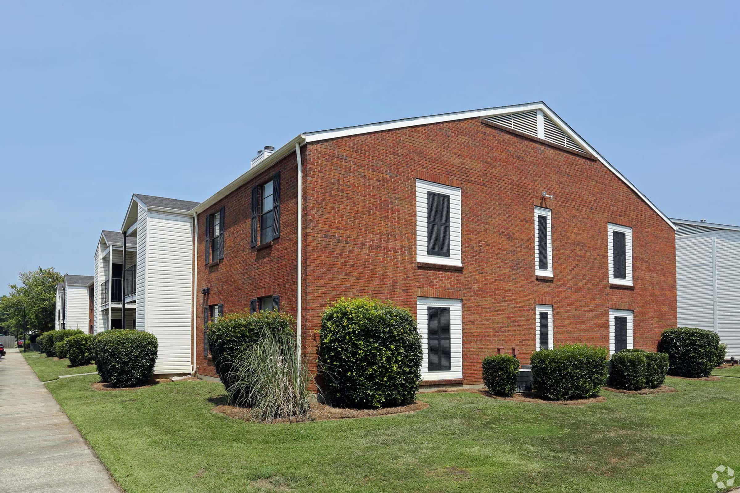 A brick apartment building with white siding, featuring windows with black shutters. The building is surrounded by neatly trimmed bushes and a well-maintained lawn. A paved walkway leads towards the entrance, under a clear blue sky.