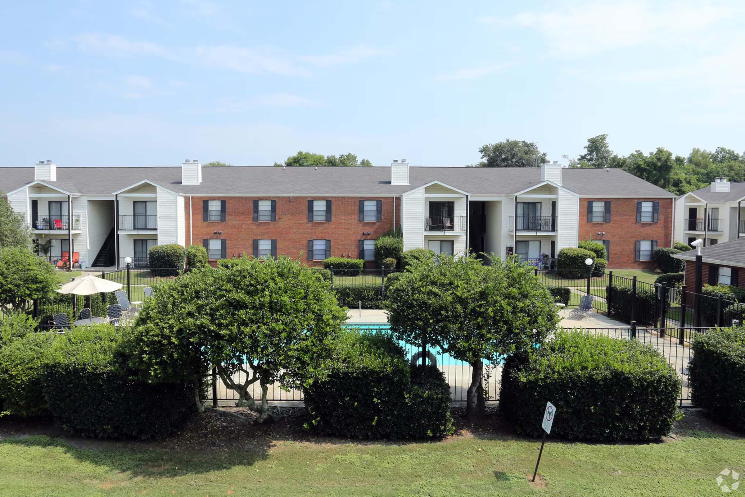 View of a landscaped apartment complex with two-story buildings featuring brick and white siding. In the foreground, neatly trimmed bushes surround a fenced swimming pool area with patio furniture. Sunny blue sky is visible above, indicating a warm day.