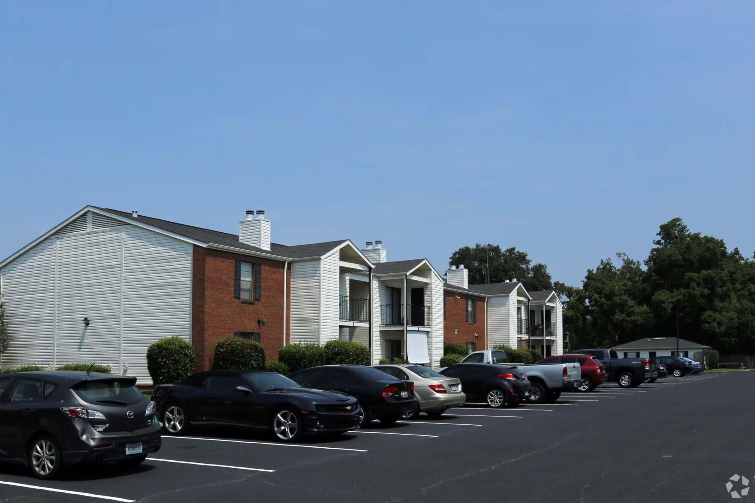 A row of multi-unit residential buildings with a mix of siding styles, set against a clear blue sky. A parking lot filled with various cars is in the foreground, with well-maintained shrubs lining the driveway. The scene conveys a suburban living environment.
