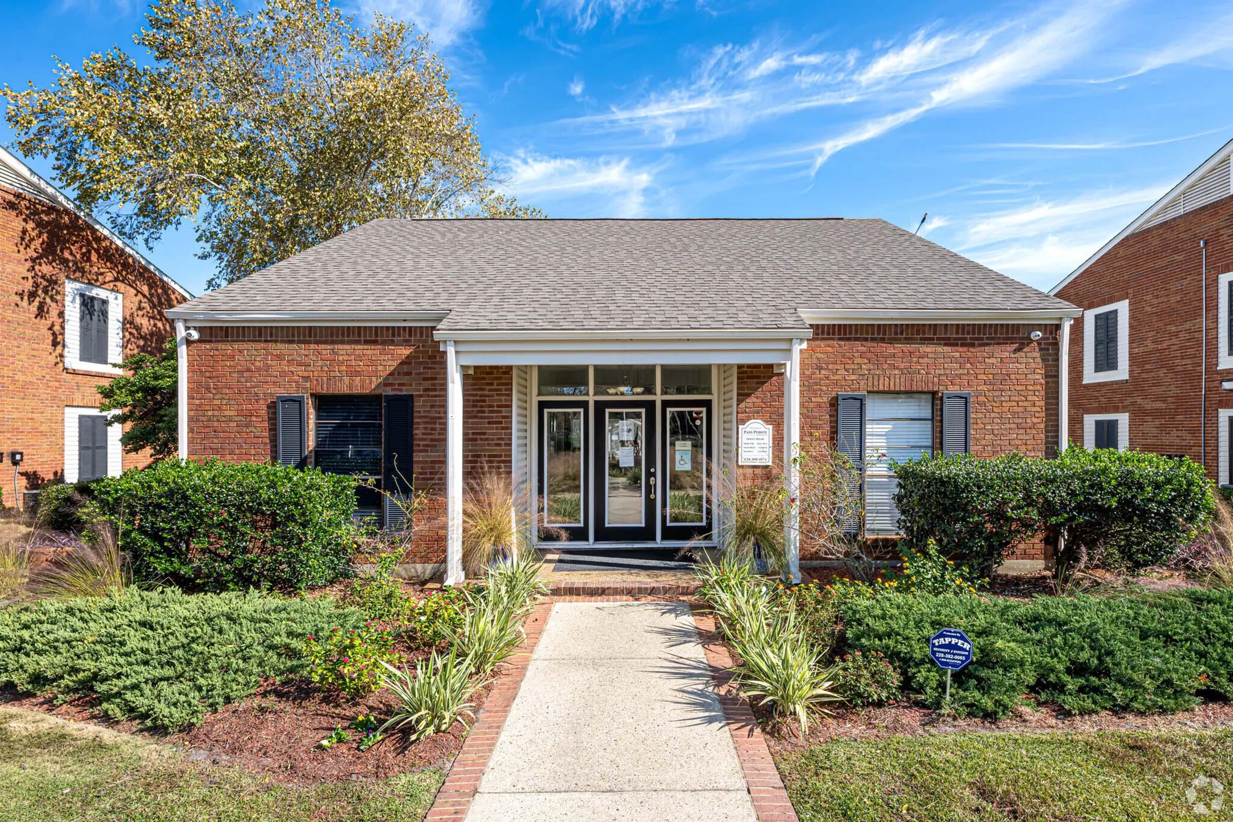 A brick building with a peaked roof, featuring a central entrance with glass doors and black shutters. Surrounding the entrance are landscaped green shrubs and plants. The building is flanked by two other structures, and a clear blue sky with scattered clouds is visible overhead.
