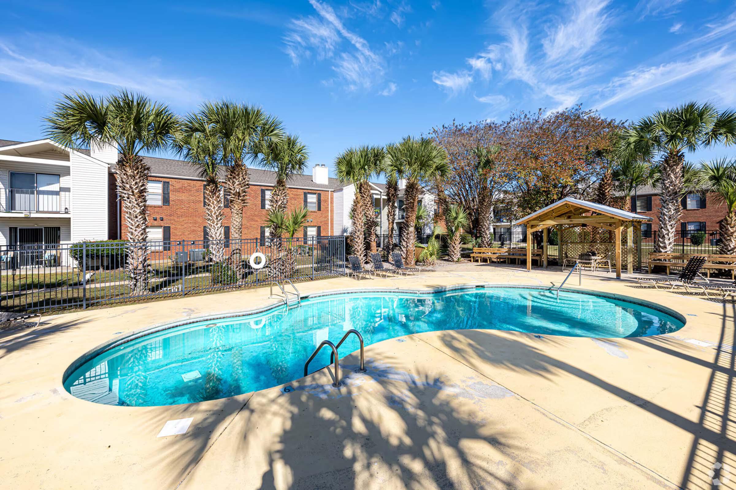 A sparkling blue swimming pool surrounded by palm trees and lounge chairs, with apartment buildings in the background. The sky is bright and clear, enhancing the inviting atmosphere of the pool area. A gazebo is also visible nearby, adding to the outdoor leisure space.