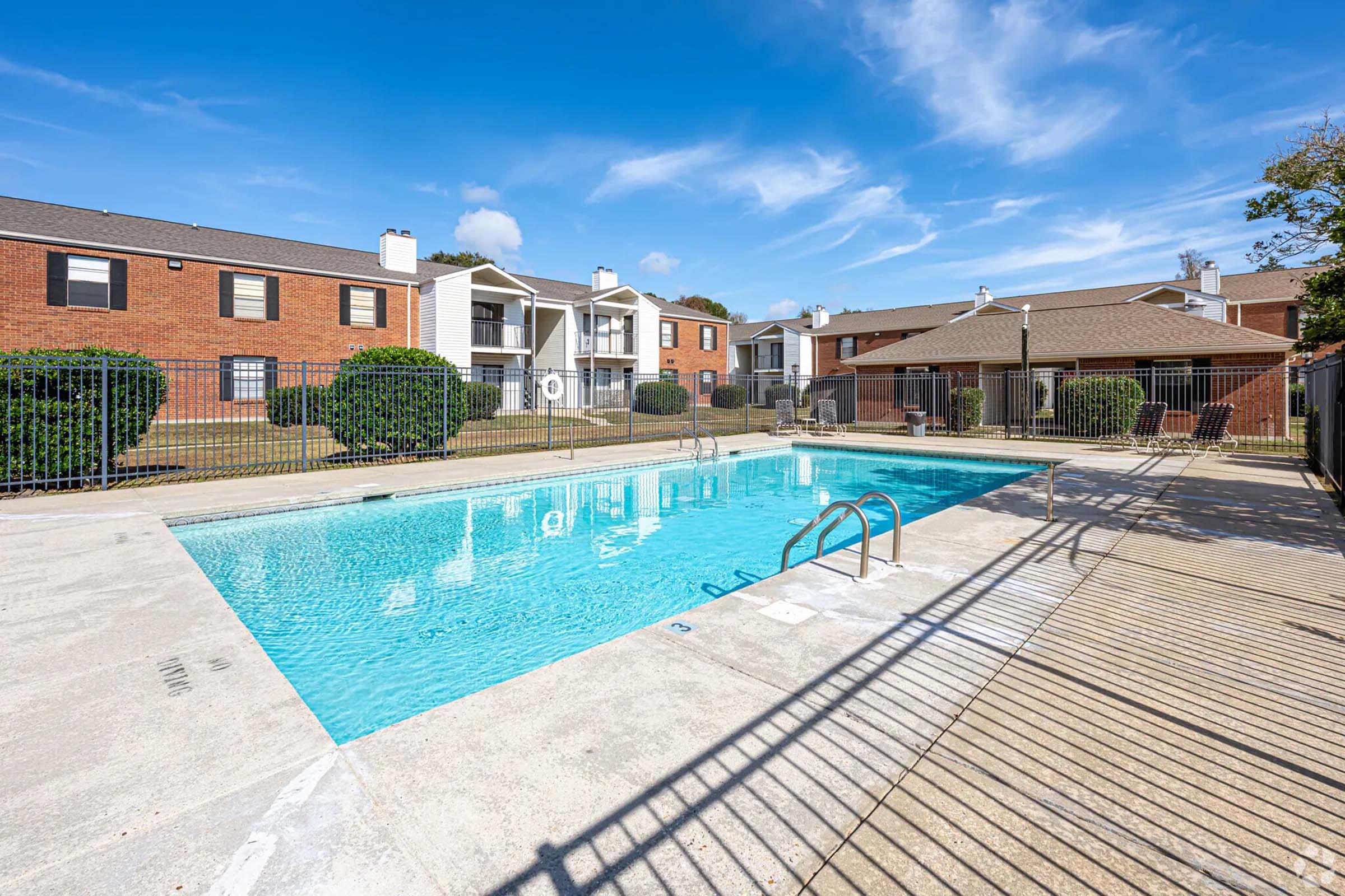 A clean, inviting outdoor swimming pool surrounded by a wooden deck, with landscaped shrubbery in the background. Several apartment buildings are visible nearby under a bright blue sky with a few clouds.