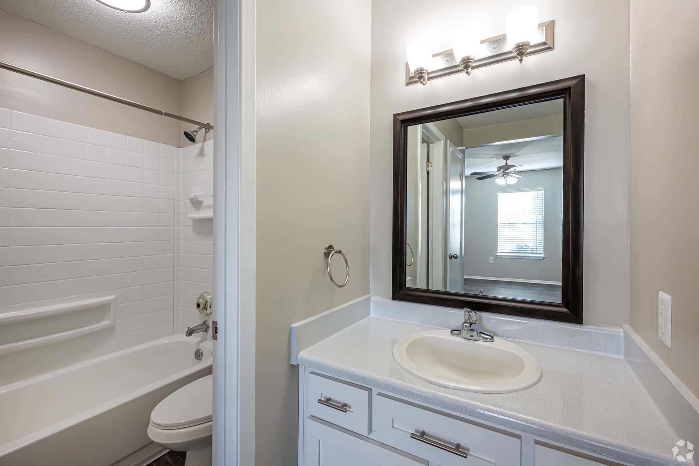 A clean, well-lit bathroom featuring a white bathtub with a shower, a modern sink with a countertop, and a large mirror framed in dark wood. The walls are painted a light color, and there’s a ceiling fan visible in the background, along with a window that lets in natural light.