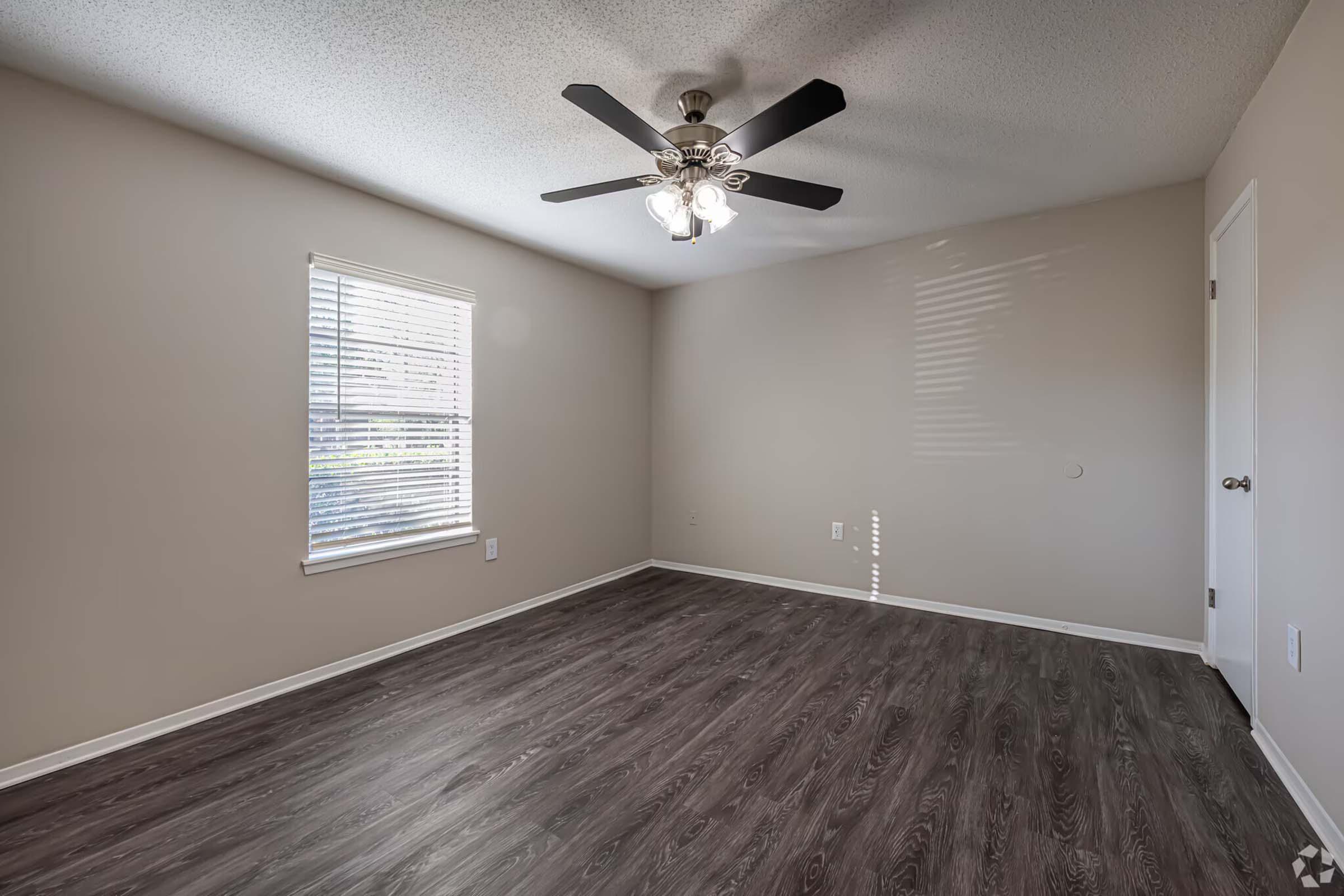 A clean, empty room featuring a ceiling fan, a window with blinds allowing natural light, and light gray walls. The floor is dark laminate, and there's a door on the right side.