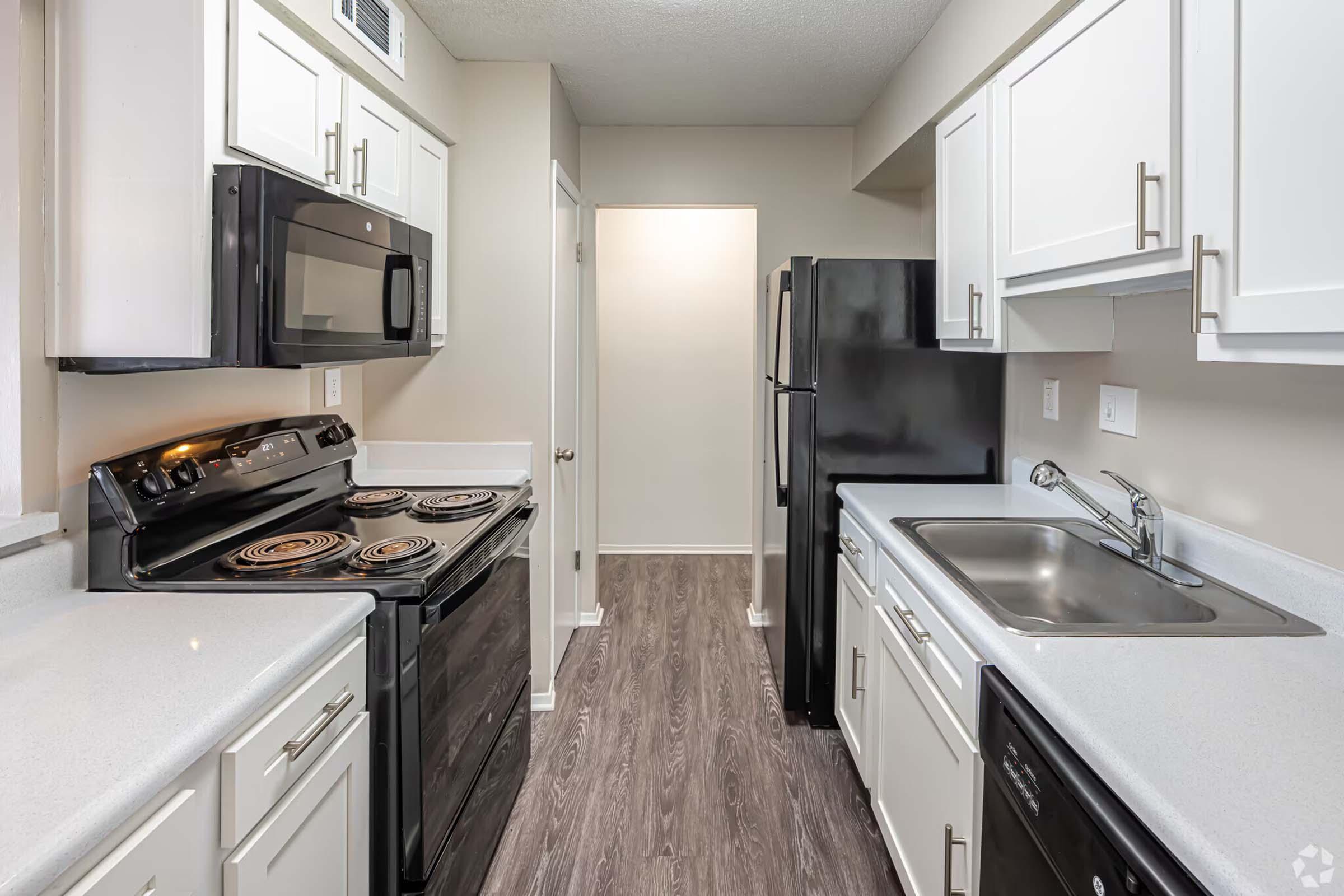 Modern kitchen featuring white cabinets, black appliances including an electric stove, microwave, and refrigerator. The countertop is light-colored, and the space is well-lit. A doorway at the back leads to another room. The flooring is a dark wood look.