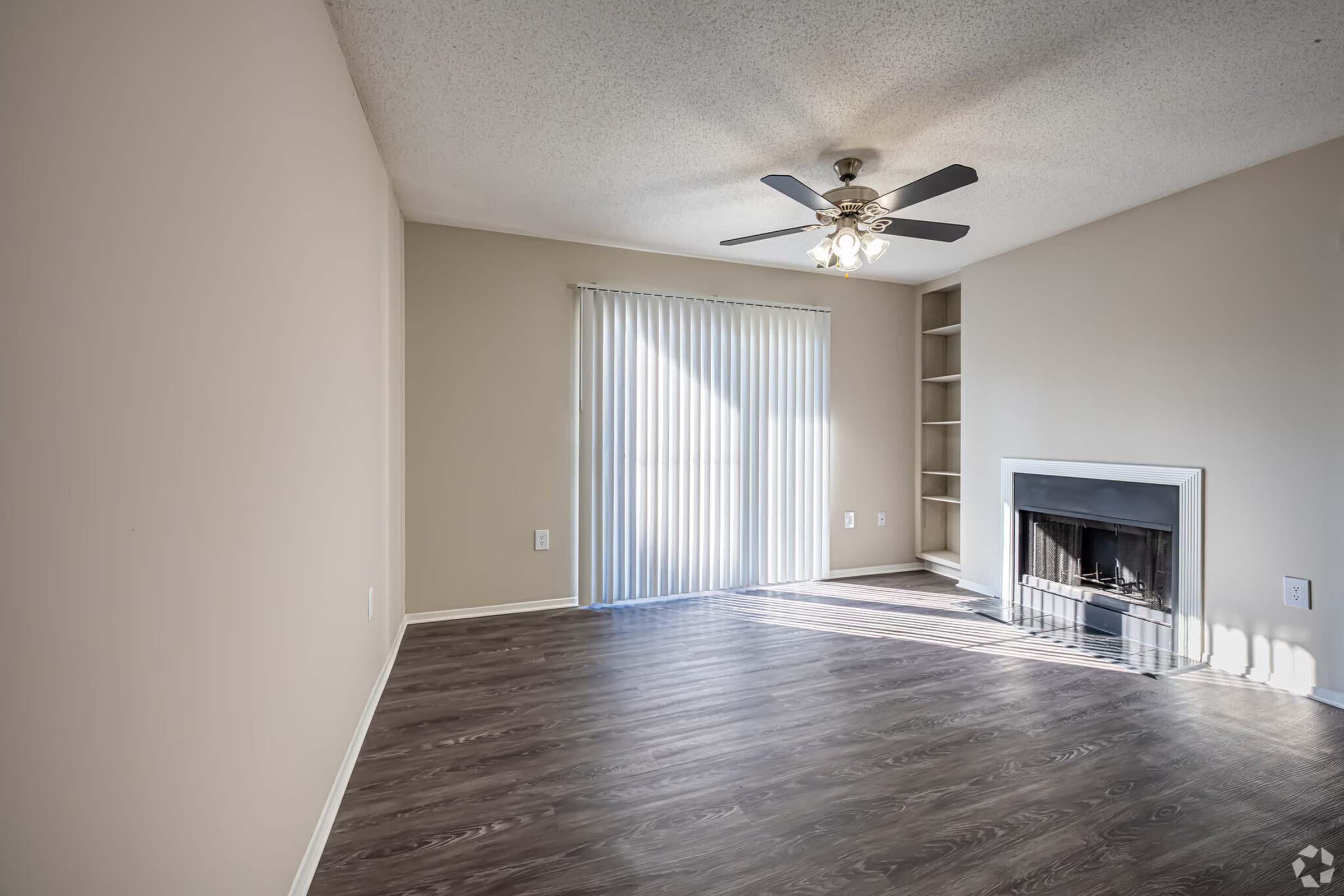 Spacious living room with wood laminate flooring, ceiling fan, and large window covered by vertical blinds. Features a neutral wall color and a traditional fireplace with a mantel. Natural light fills the room, creating a warm and inviting atmosphere.