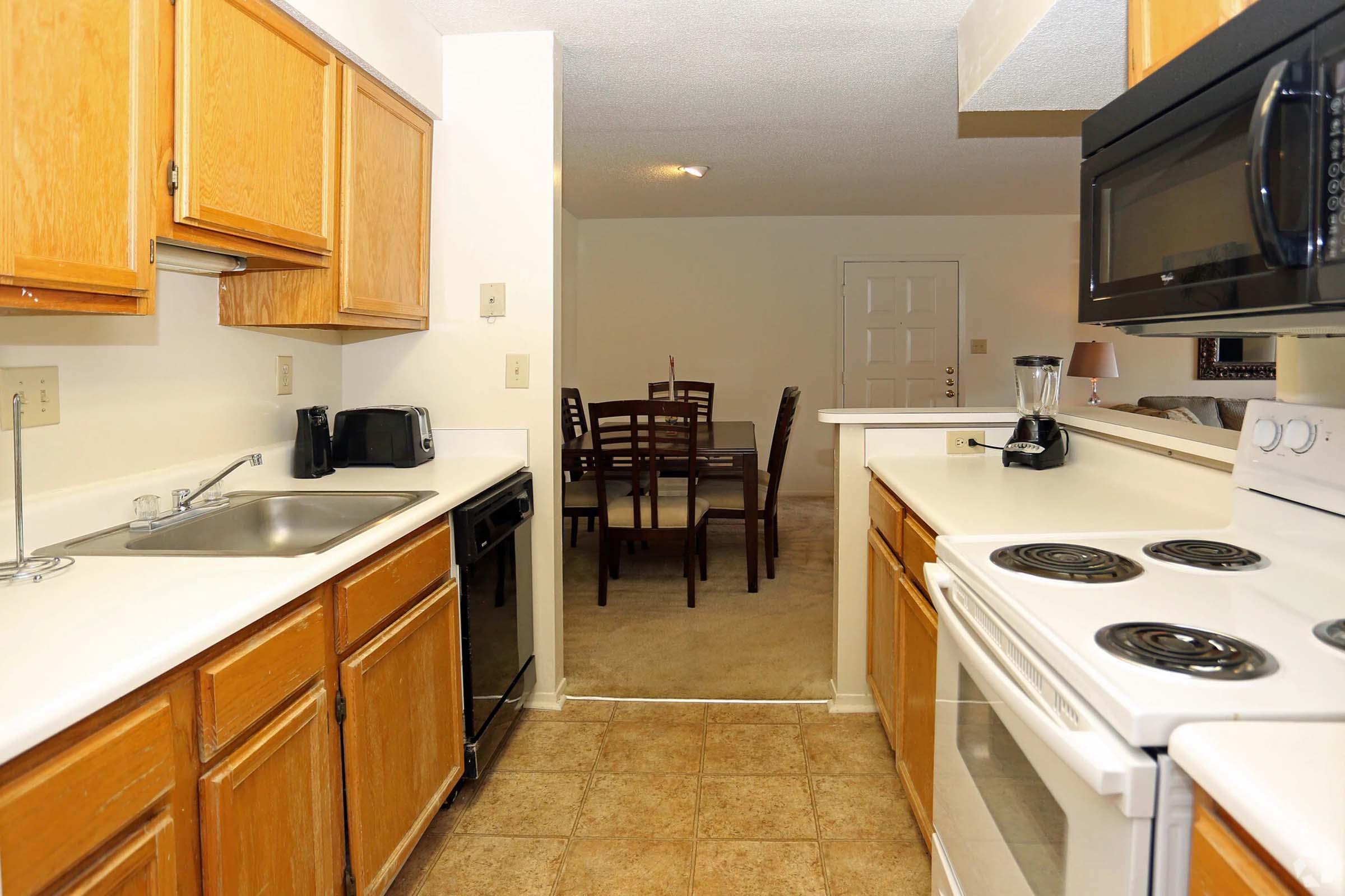 A well-lit kitchen featuring light wood cabinetry, a white countertop, a stainless steel sink, and a black microwave. The kitchen includes a dishwasher and a stove with an oven. In the background, a dining area with a wooden table and chairs is visible, along with carpeted flooring and a doorway leading to another room.