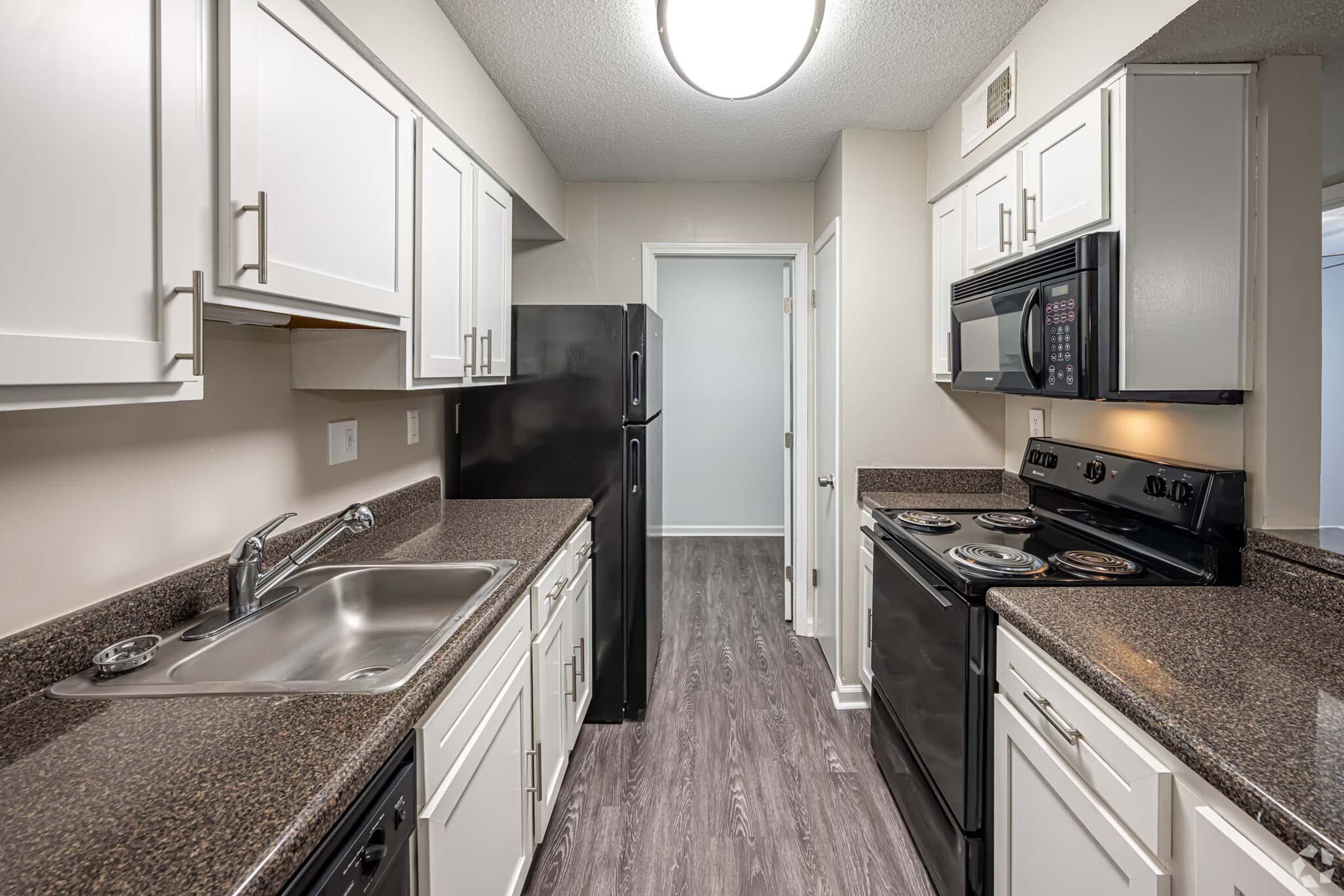 A modern kitchen featuring dark countertops, white cabinets, and stainless steel appliances. The space includes a sink, stove, microwave, and refrigerator. The flooring is a wood-like laminate, and there is an open doorway leading to another room. The overall aesthetic is clean and contemporary.