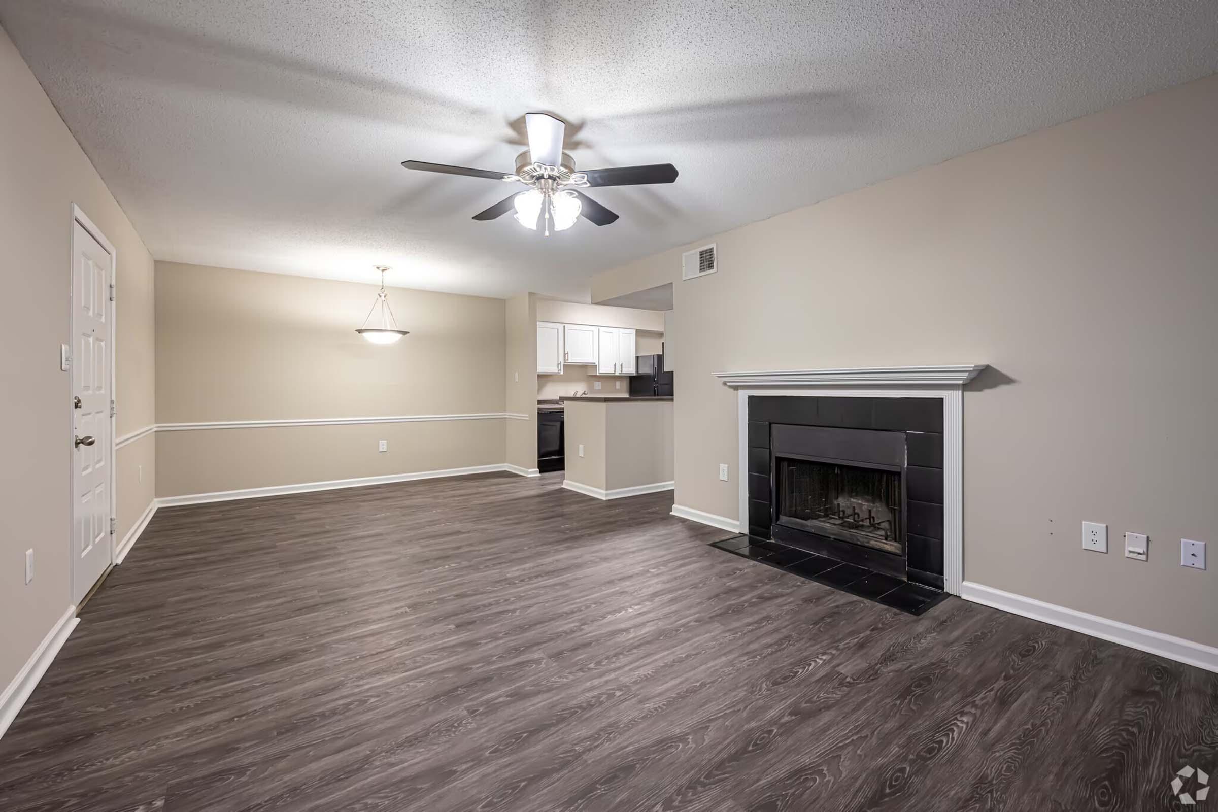 Spacious interior of a living room featuring light-colored walls, a ceiling fan, and laminate flooring. The room includes a modern fireplace with a black surround and an open layout leading to a kitchen area, with a doorway visible on the left. Natural light from a nearby fixture enhances the inviting atmosphere.