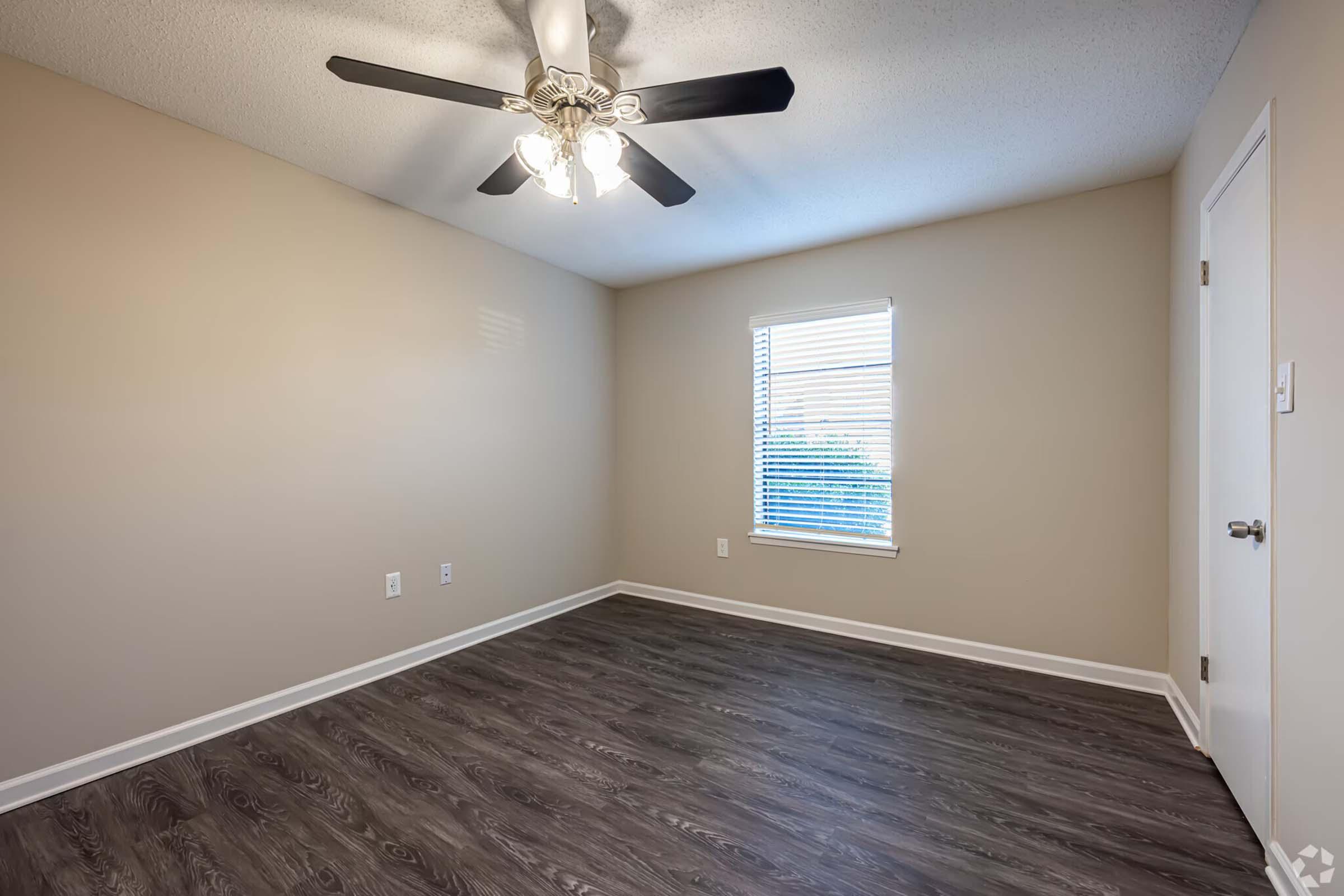 A clean, empty room featuring a light-colored wall, a ceiling fan with lights, a window with blinds allowing natural light, and dark laminate flooring. The room has no furniture, emphasizing its spaciousness.