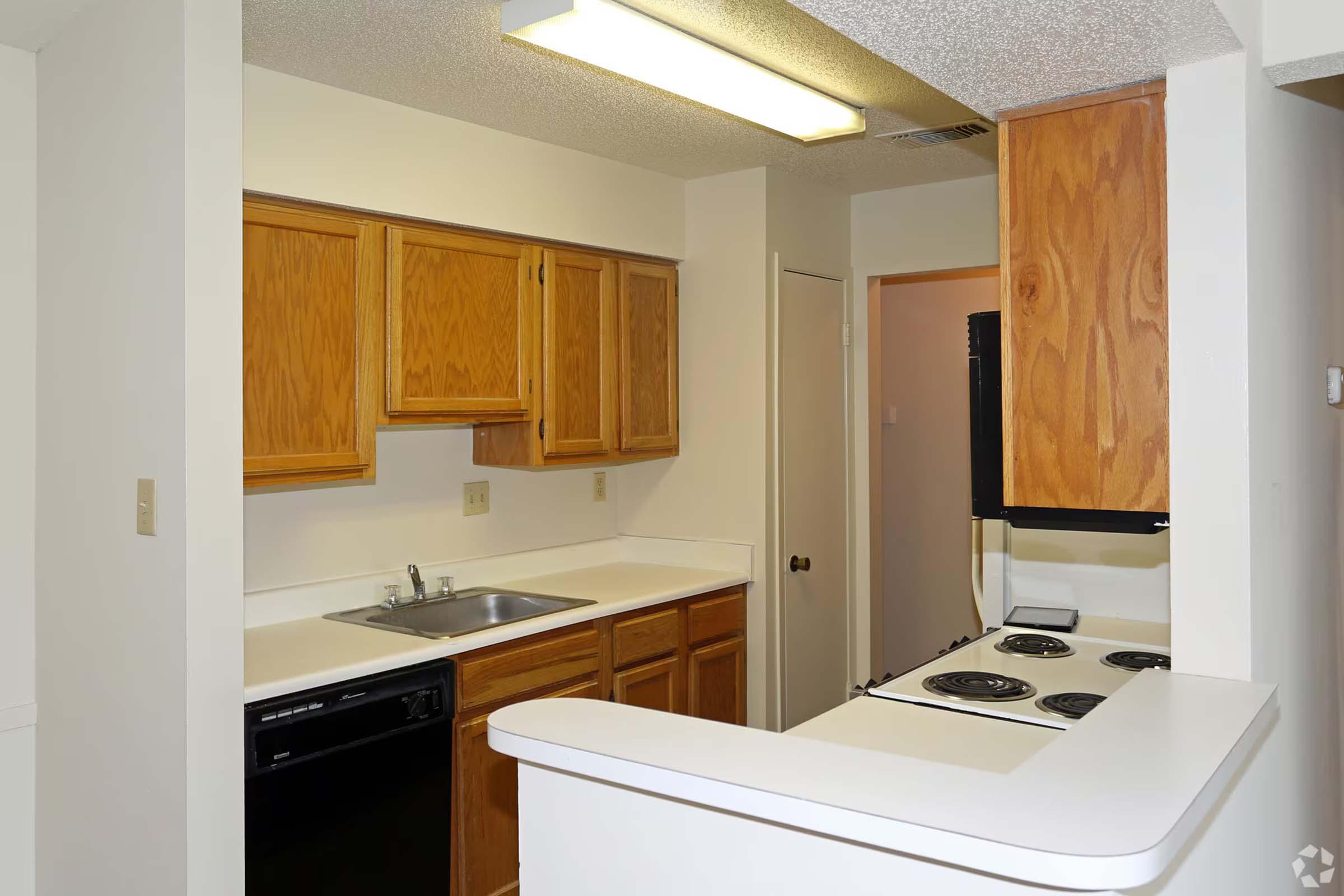 A compact kitchen with wooden cabinets, a stainless steel sink, a black dishwasher, and a stove with four burners. The layout features a bar counter that separates the kitchen from a living space, with a light fixture above and a neutral color scheme throughout.