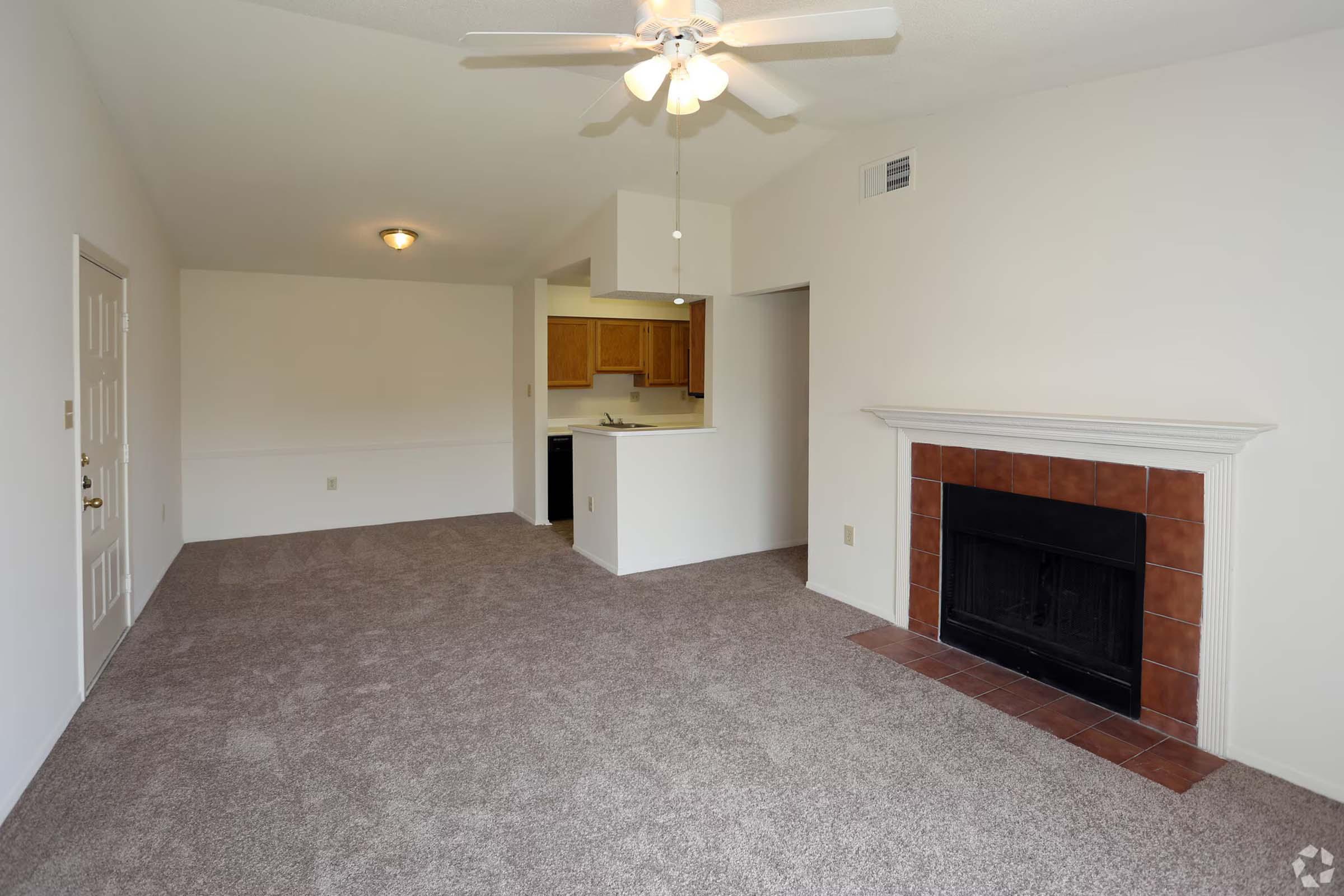 A spacious living room featuring beige carpet, a ceiling fan, and a fireplace with a tiled hearth. To the left, there is a door, and in the background, an open kitchen area with wooden cabinets is visible. The walls are painted a light color, enhancing the room's brightness.
