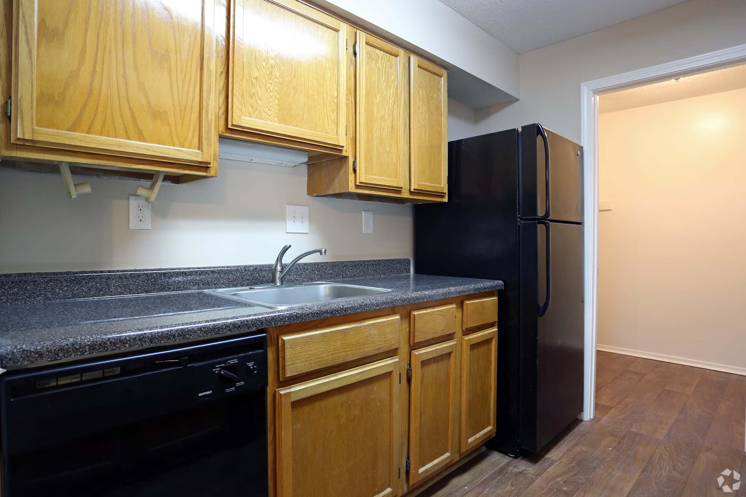 A kitchen featuring wooden cabinetry, a black refrigerator, a sink with a faucet, and a black dishwasher. The countertop is dark, and the floor has a wood-like finish. A doorway leads to another room in the background.