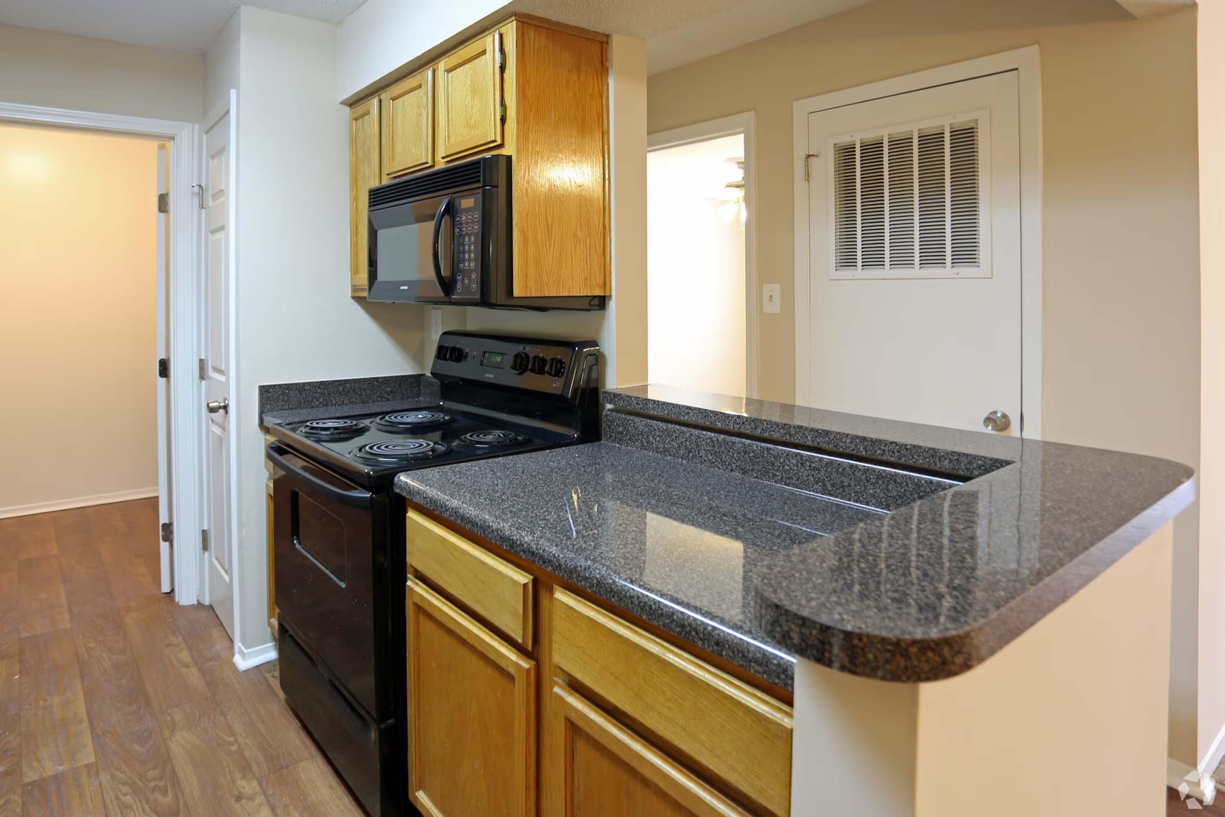 A small kitchen featuring wooden cabinets, a black stove, and a microwave mounted above it. The countertops are dark granite, and the space has light-colored walls and wooden flooring. A doorway leads to another room, indicating an open layout.