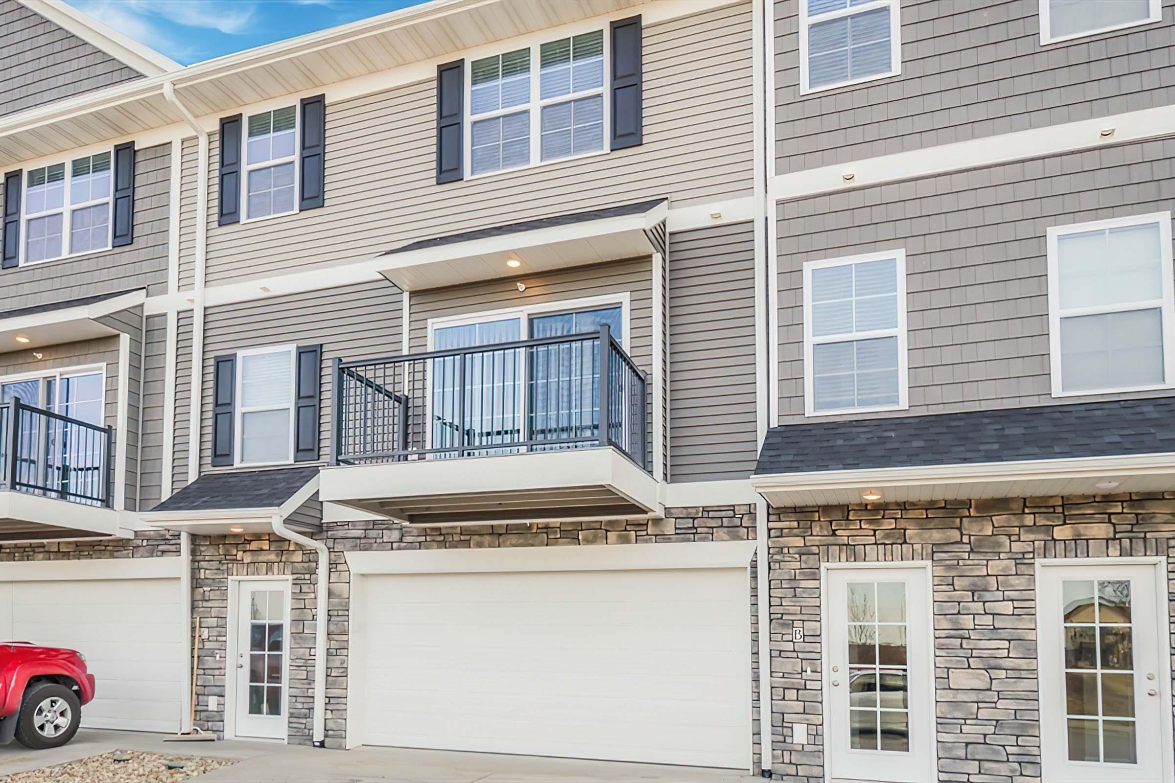 Exterior of a modern townhouse featuring a stone and siding facade. The front includes a garage door, a small balcony on the second floor, and large windows. A red vehicle is parked in front. The overall design is contemporary with a neutral color palette. Blue sky in the background.