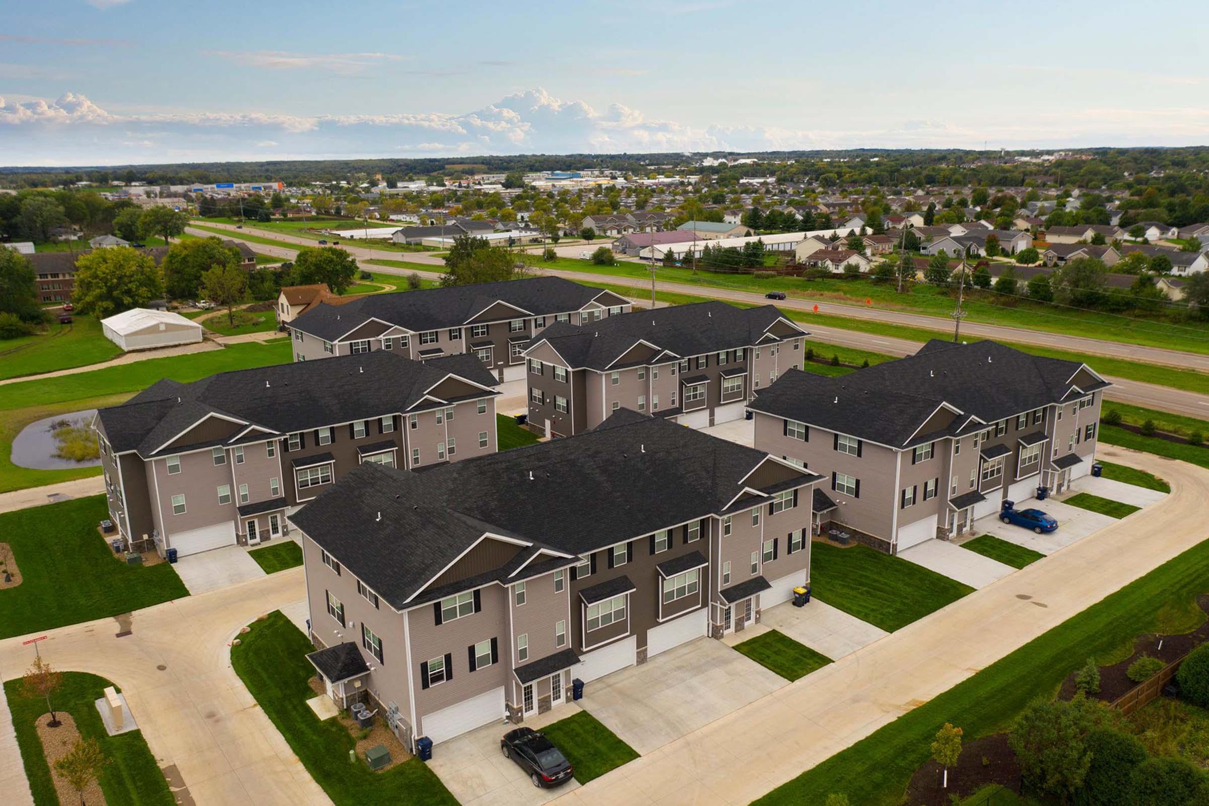 Aerial view of a residential area featuring several multi-unit buildings with gray siding and dark roofs. Well-maintained landscaping surrounds the buildings, and there are visible parking spaces. In the background, a highway and other residential neighborhoods are seen, along with a few clouds in the sky.