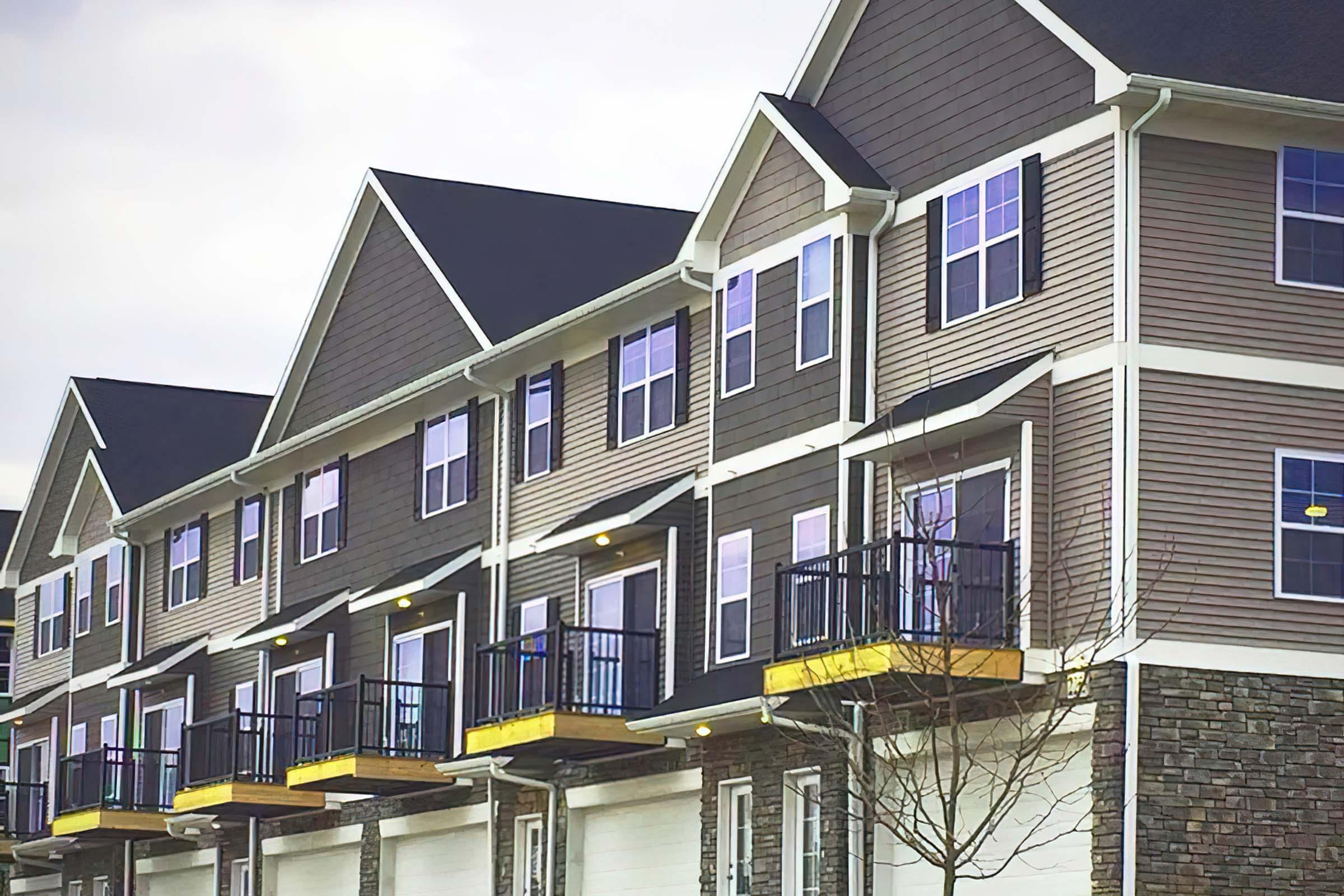 A row of modern townhouses featuring multiple stories, large windows, and balcony space. The buildings have a contemporary design with a combination of siding materials and stone accents. The overcast sky adds a slight gray hue to the scene.
