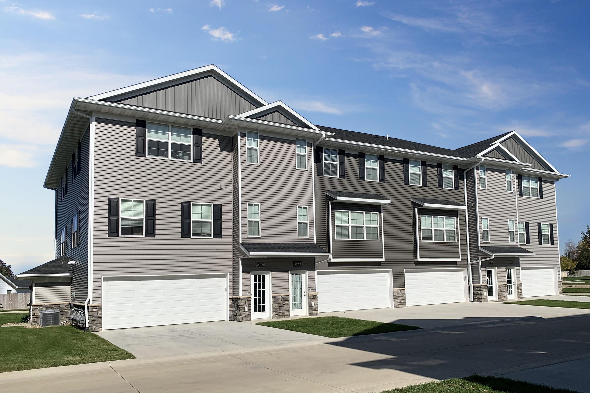 A modern multi-unit residential building featuring three stories, with a light gray exterior and dark accents. The structure includes multiple large windows and garages on the ground floor. The setting is outdoors with a clear blue sky and green grass in the foreground.