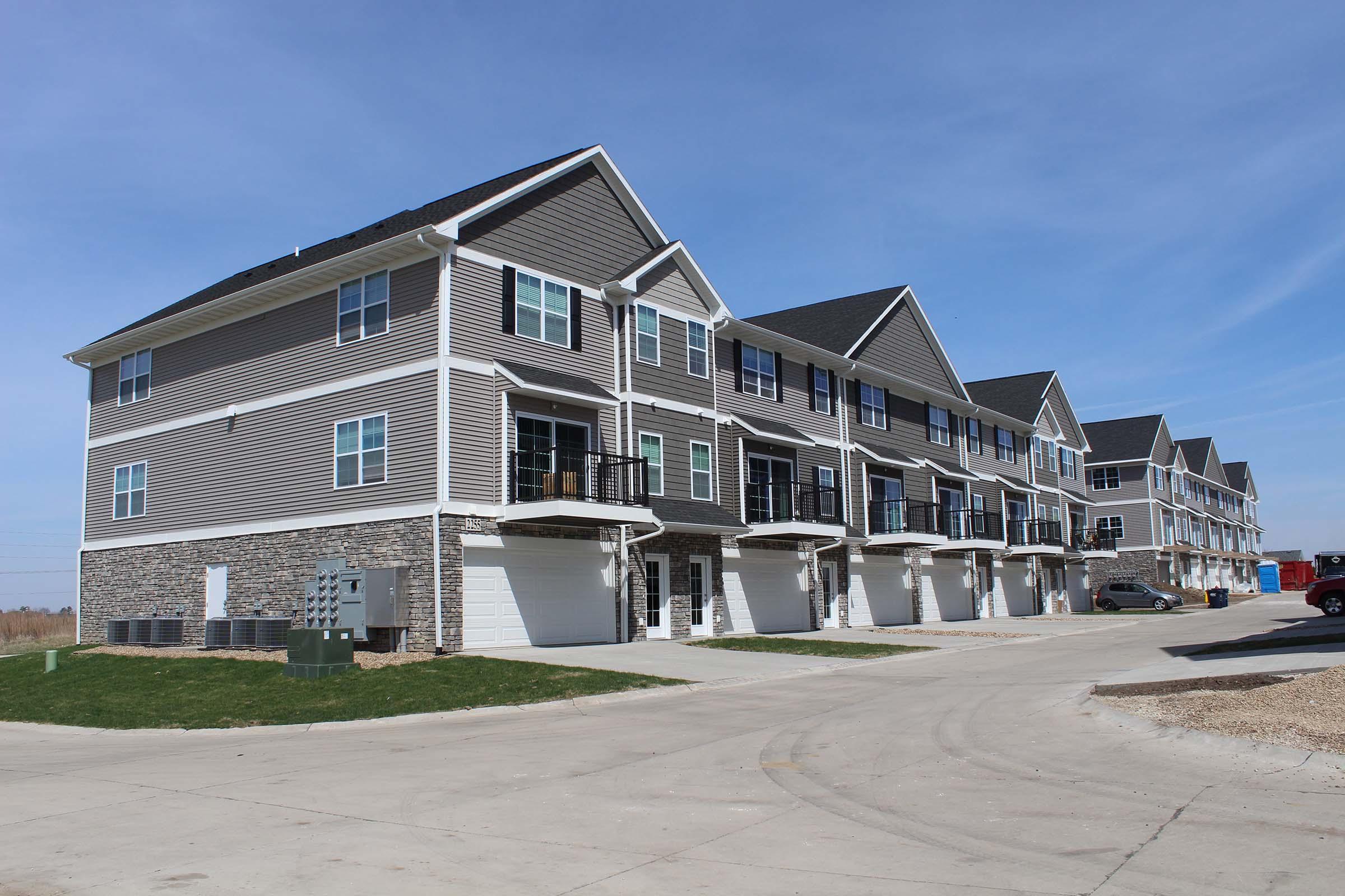 A row of modern townhouses featuring gray siding, stone accents, and multiple balconies. The building has several garage doors on the ground level, and a paved driveway runs alongside. The sky is clear and blue, creating a bright atmosphere in the neighborhood.