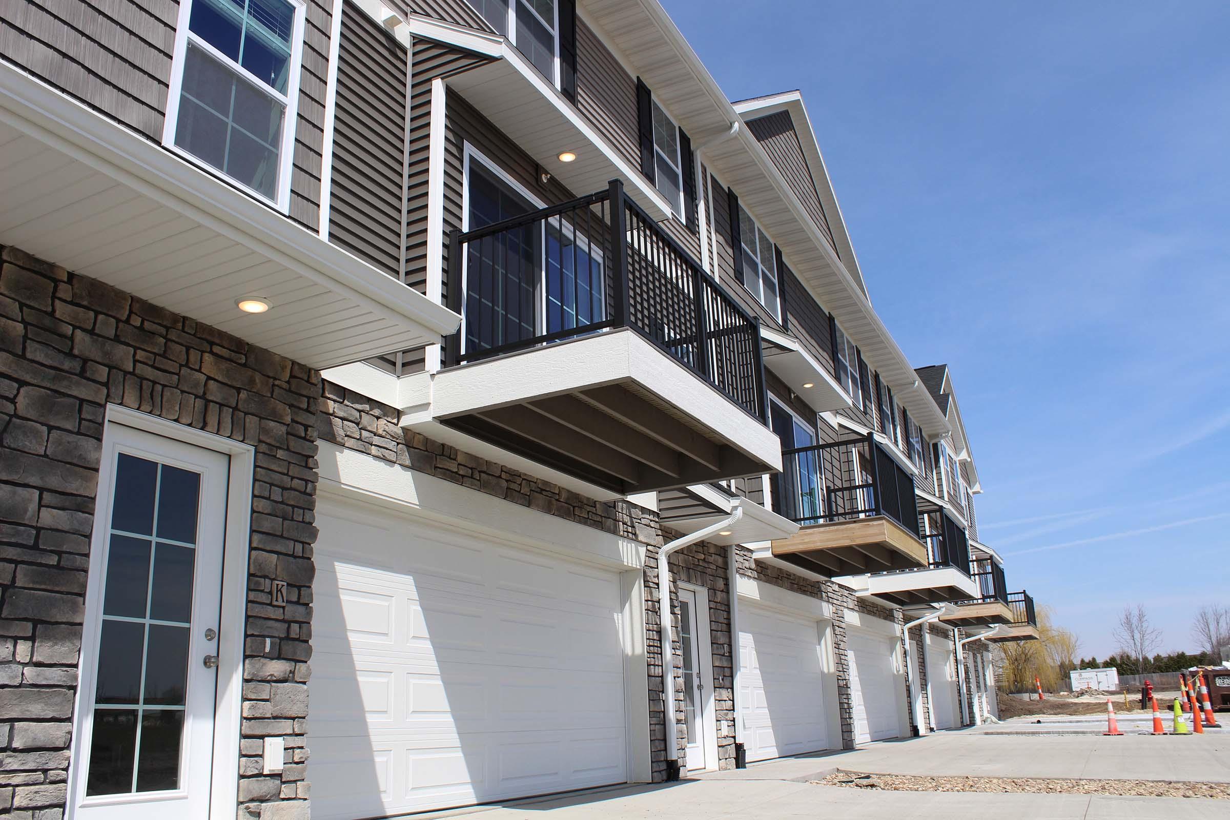 Contemporary townhouse-style buildings with stone and siding exteriors. Each unit features a balcony, a garage door, and large windows. The sky is clear with a few clouds, and the surrounding area is neatly landscaped with gravel.