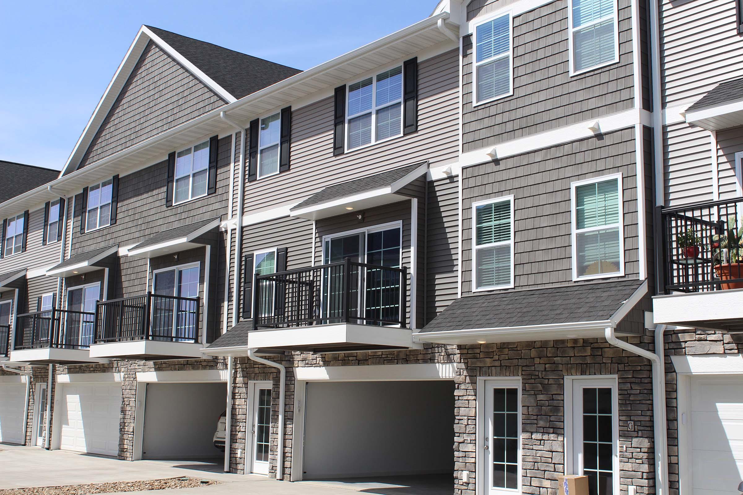 Row of modern townhouses featuring stone and siding exteriors, with balconies on the upper level and attached garages below. The buildings have multiple windows and are set against a clear blue sky.