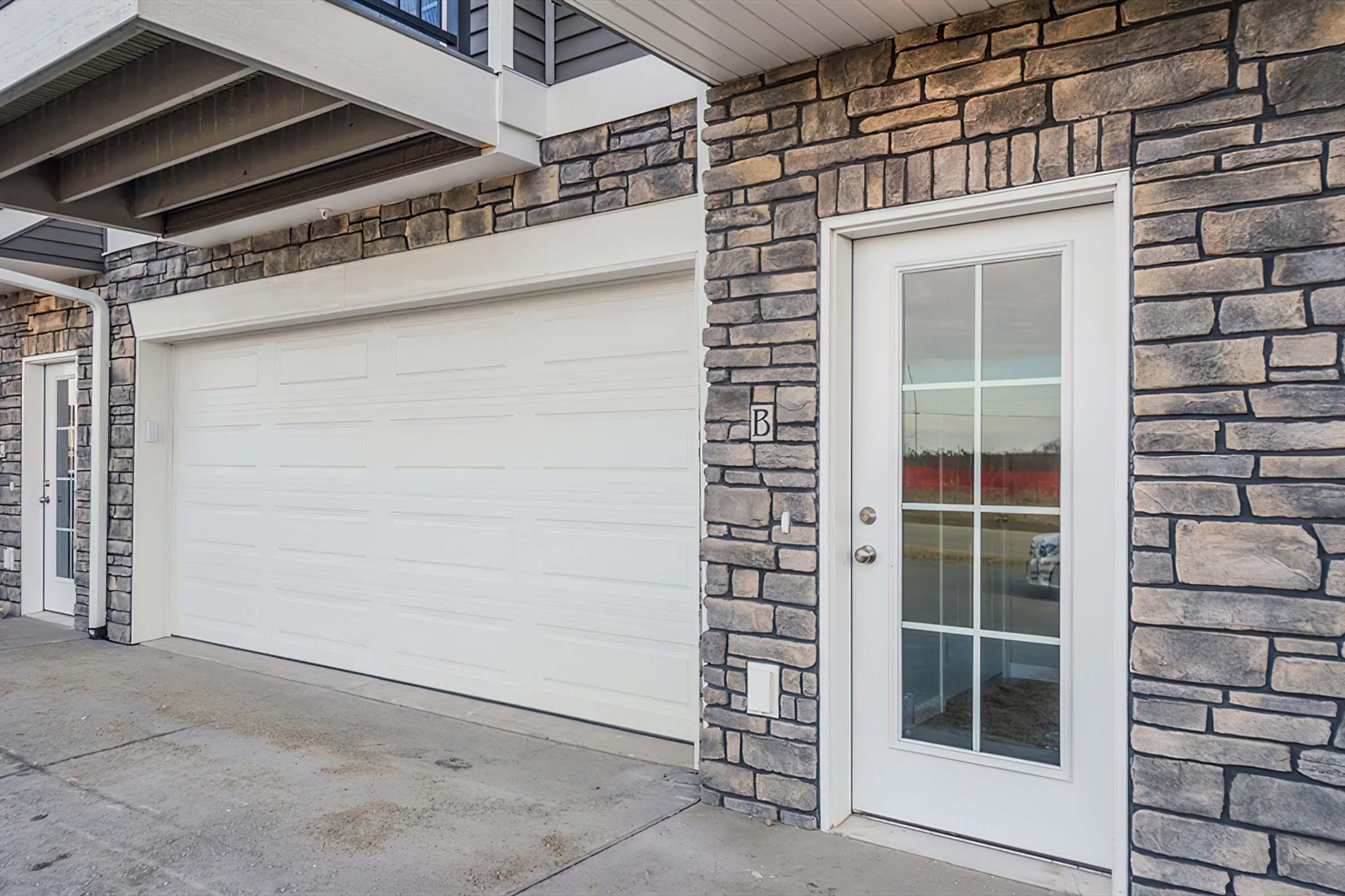 A garage door and a front door are situated beside each other, featuring a stone-accented wall. The garage door is white and large, while the front door has a glass panel and is located on a concrete walkway. The surroundings reveal a hint of greenery and a road in the background.