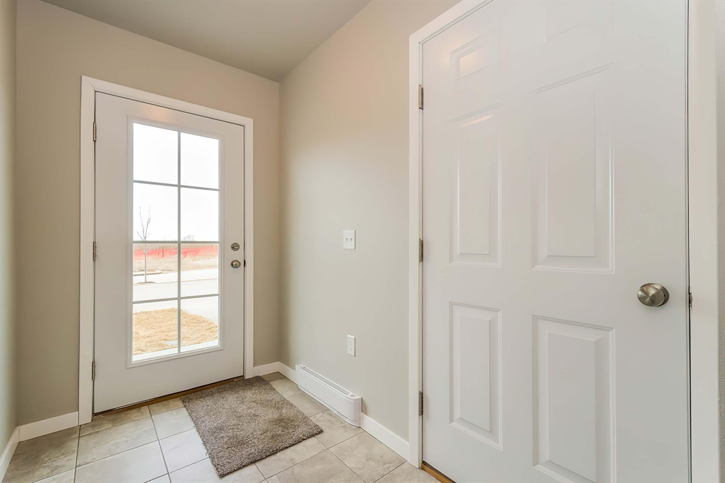 A well-lit entryway featuring a white door with glass panels and a light gray wall. There's a small welcome mat on the tile floor and a blank wall opposite the door. Natural light comes in through the window, illuminating the space.