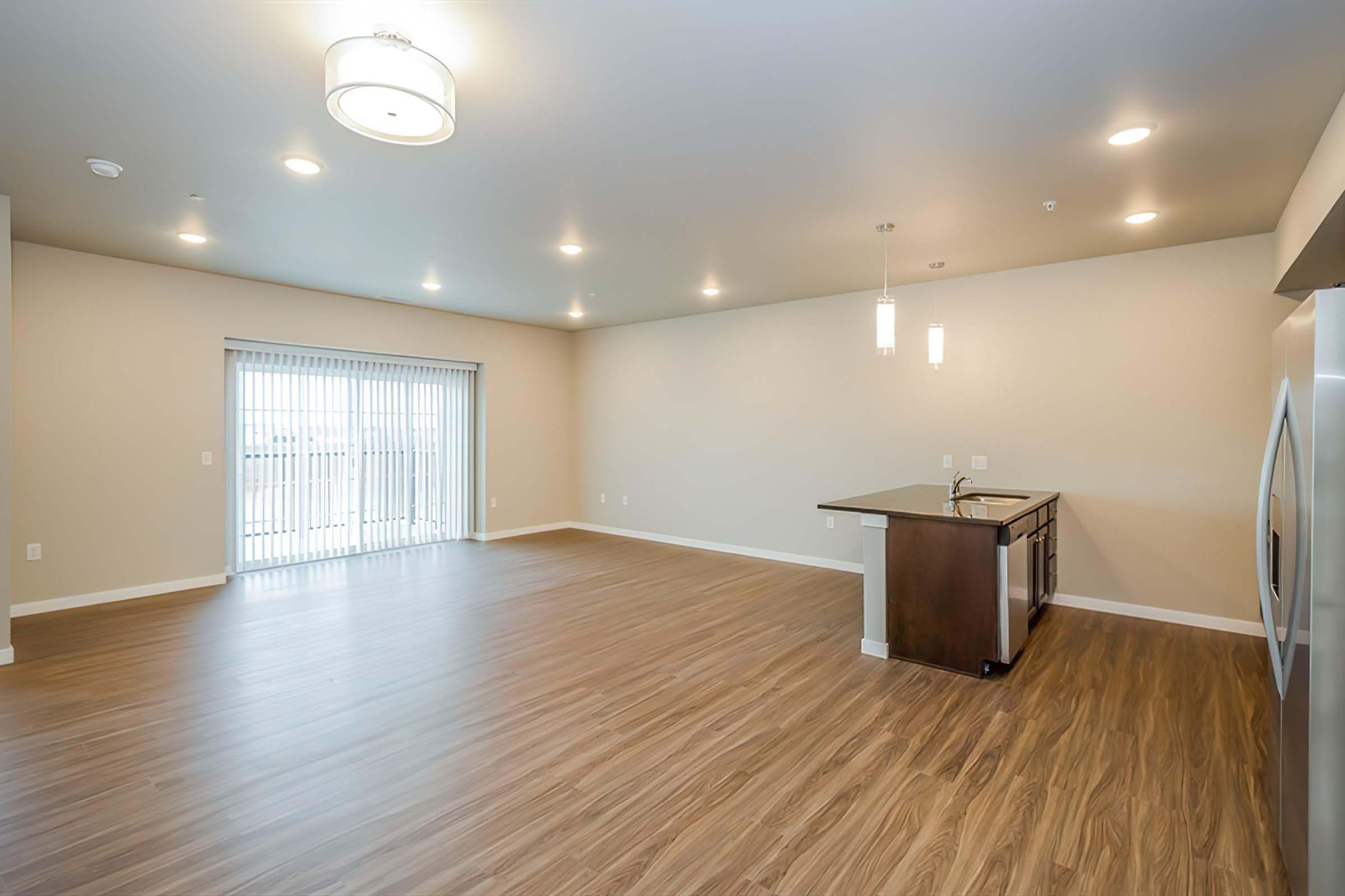 Spacious, modern living room with wood-style flooring, large windows offering natural light, and a kitchen area featuring a dark wood breakfast bar and stainless steel appliances. The walls are painted in a neutral tone, enhancing the bright and airy atmosphere. Ideal for relaxation and entertainment.